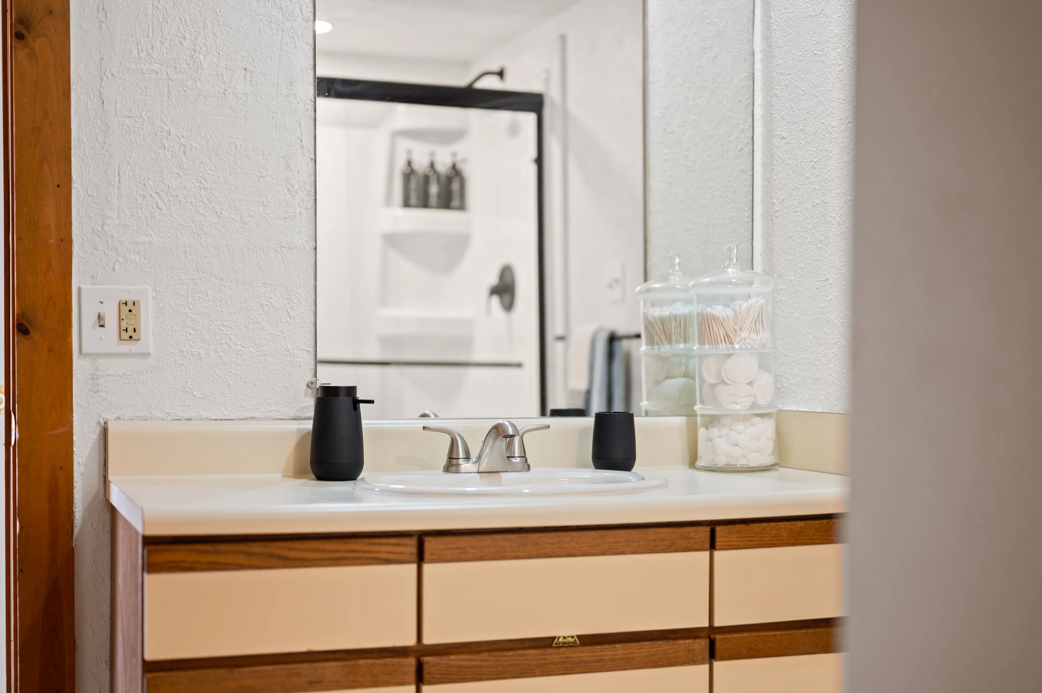 Modern bathroom vanity with a white countertop, wooden cabinets, and a mirror reflecting a glass-door shower; assorted toiletry containers arranged on the countertop.