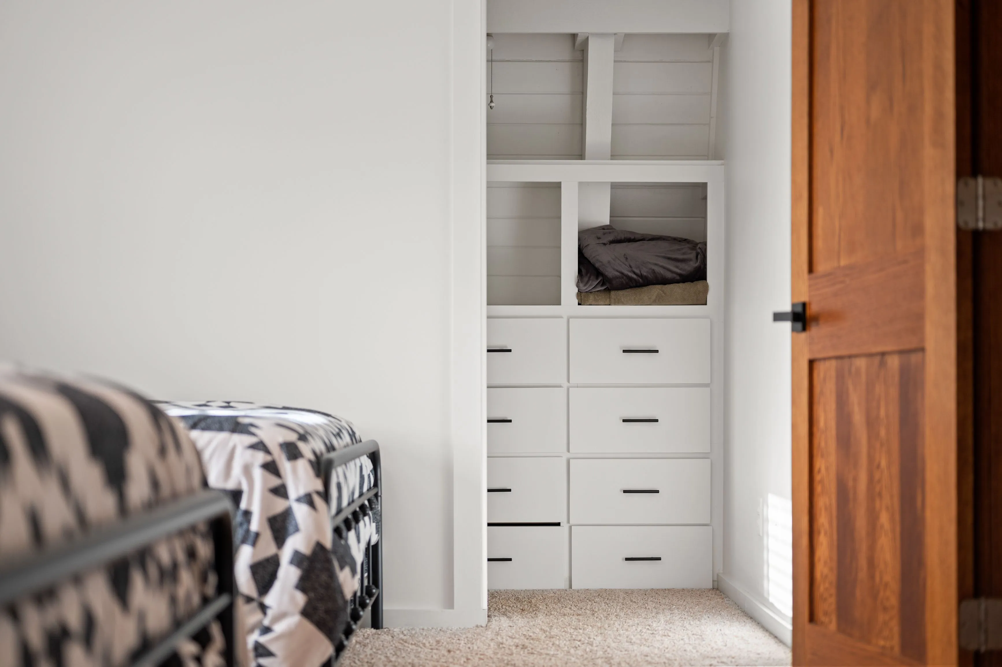 A modern bedroom with an open closet featuring built-in white drawers and shelves with a folded blanket, partial view of a patterned bedspread, and a wooden door ajar to the right.
