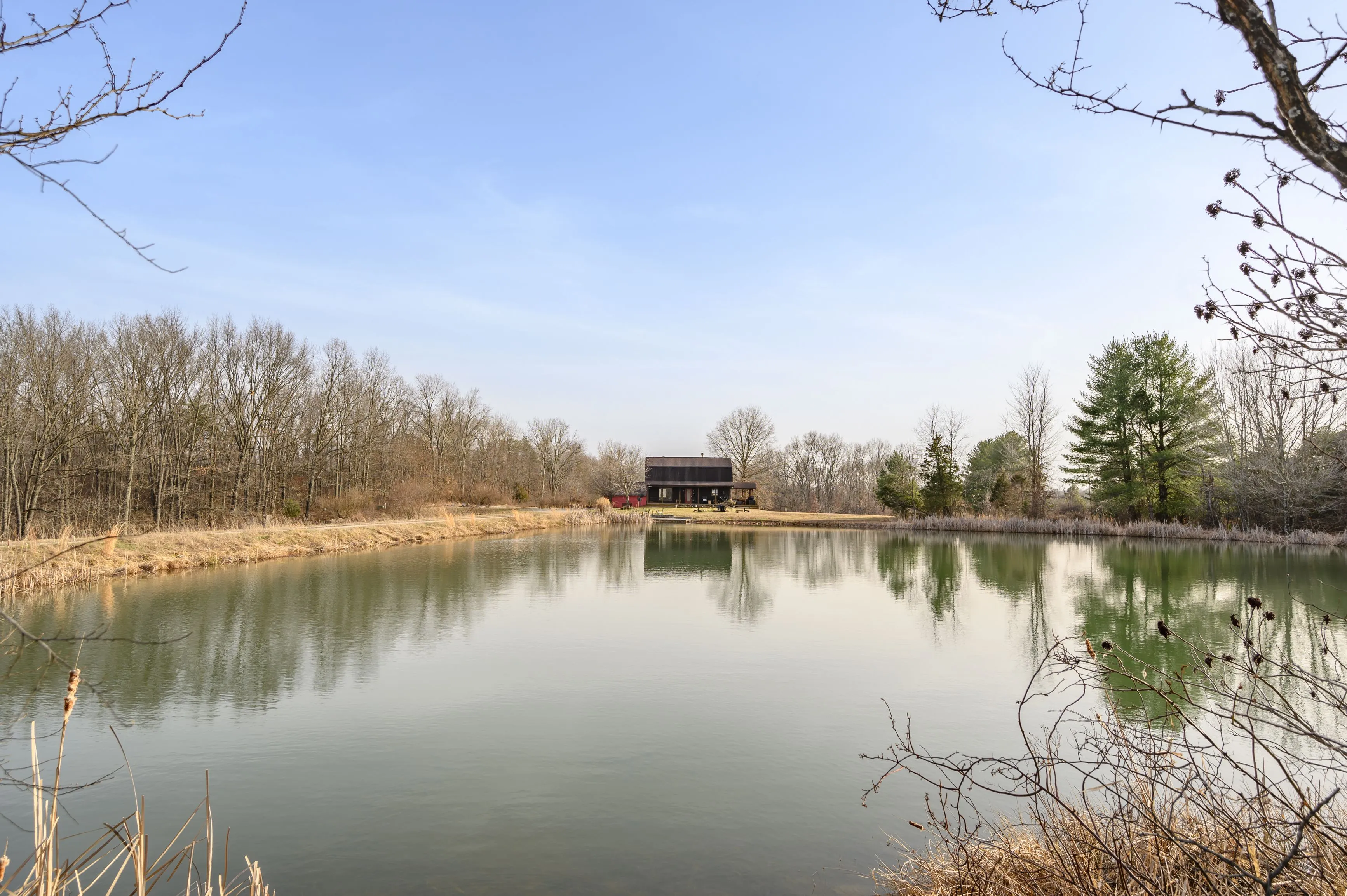 Tranquil country landscape featuring a pond with a reflection of the sky and trees, framed by bare branches in the foreground and a wooden house on the far shore.