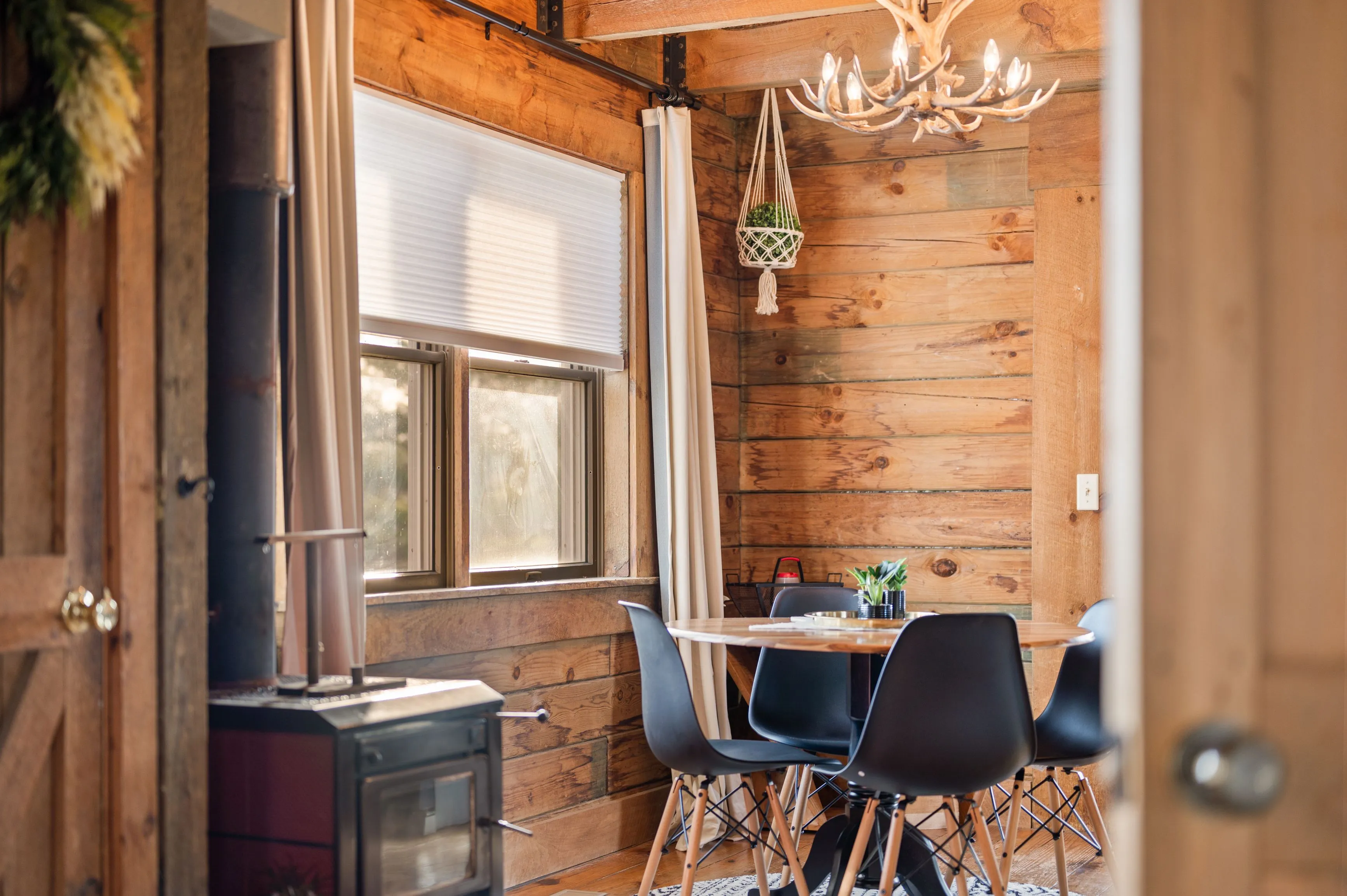 Cozy cabin interior with wooden walls, a small dining table and chairs, antler chandelier, and a wood-burning stove.