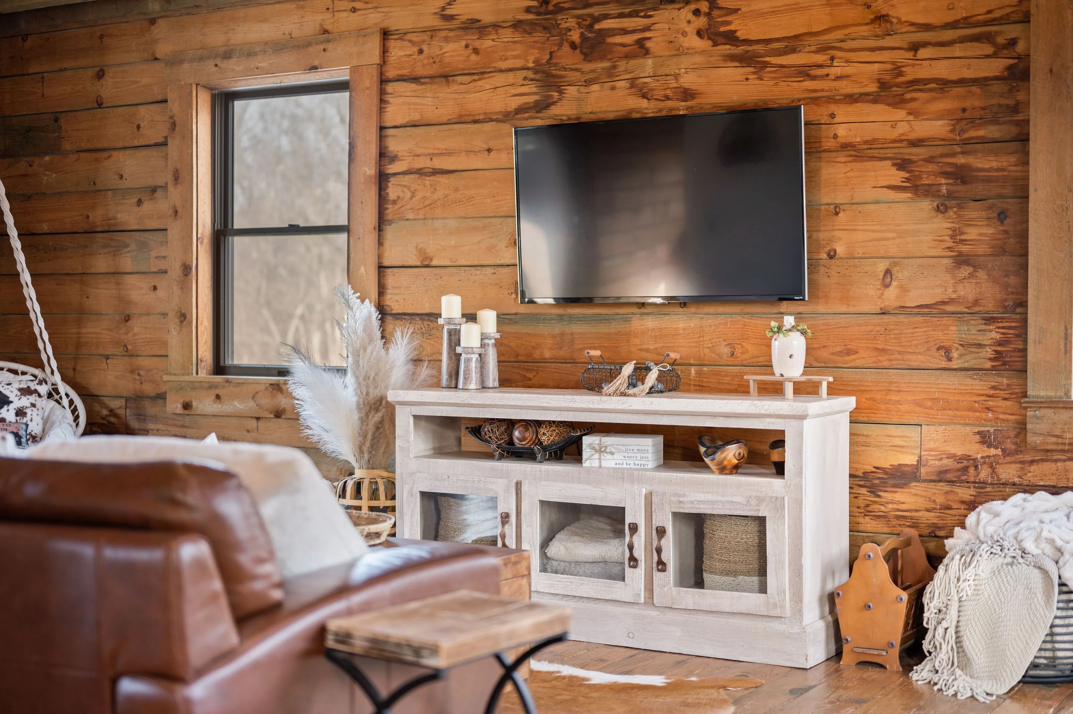 Cozy living room with rustic wooden interior, featuring a flat-screen TV mounted on the wall above a decorative console table, with a leather sofa and cowhide rug in the foreground.