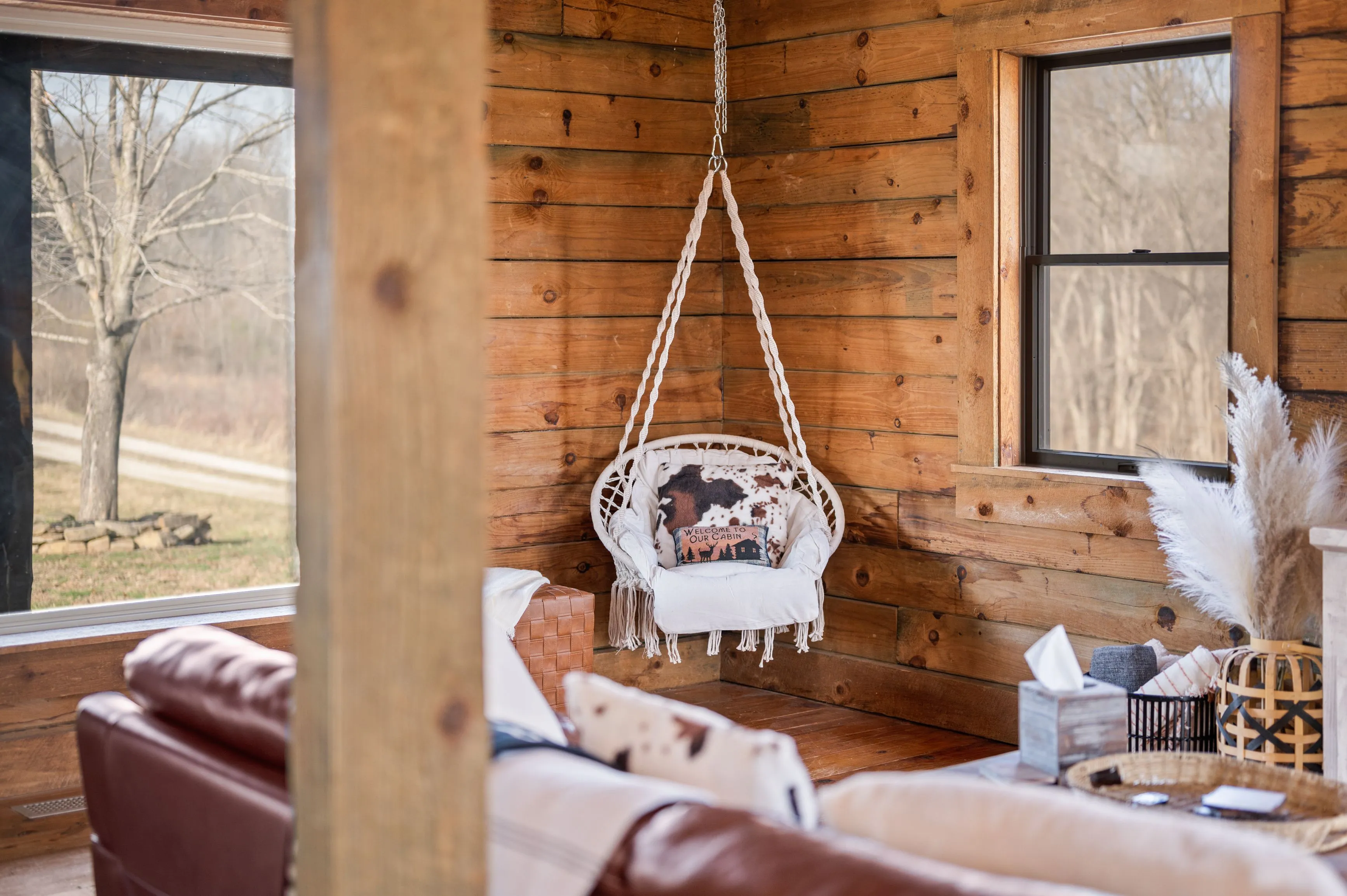 Cozy wooden cabin interior with hanging swing chair, plush cushions, and a view of outside trees through window.