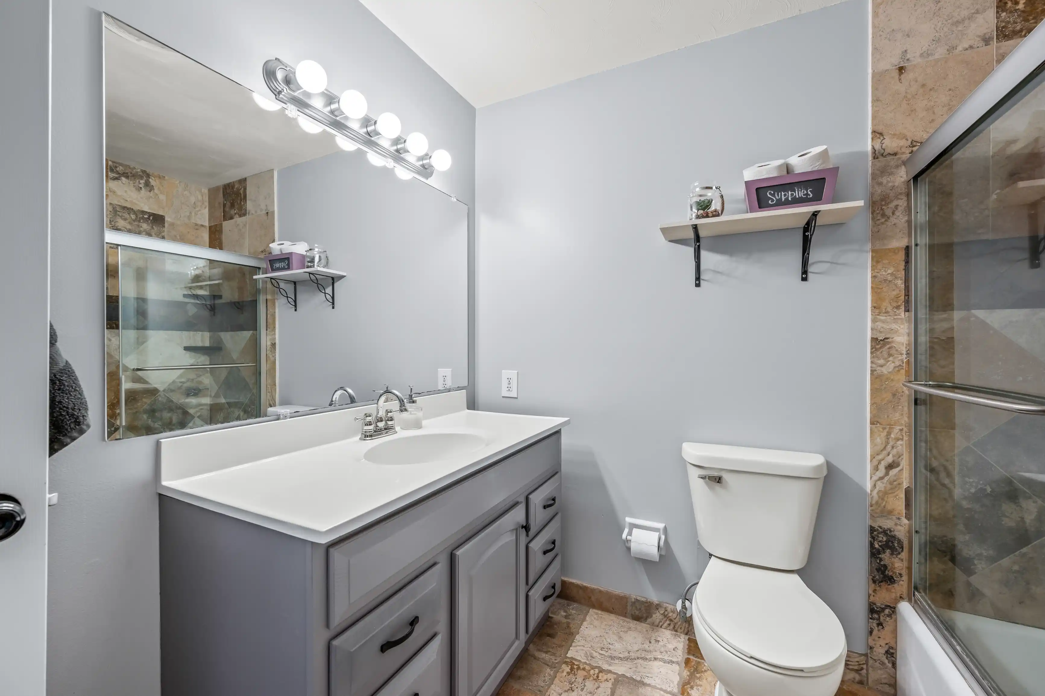 A modern bathroom with a large mirror lit by a row of lights above, a gray vanity with a white countertop and sink, a toilet to the right, and a glass-enclosed shower with stone tile walls. There is also a wooden shelf labeled 'Supplies' on the wall.