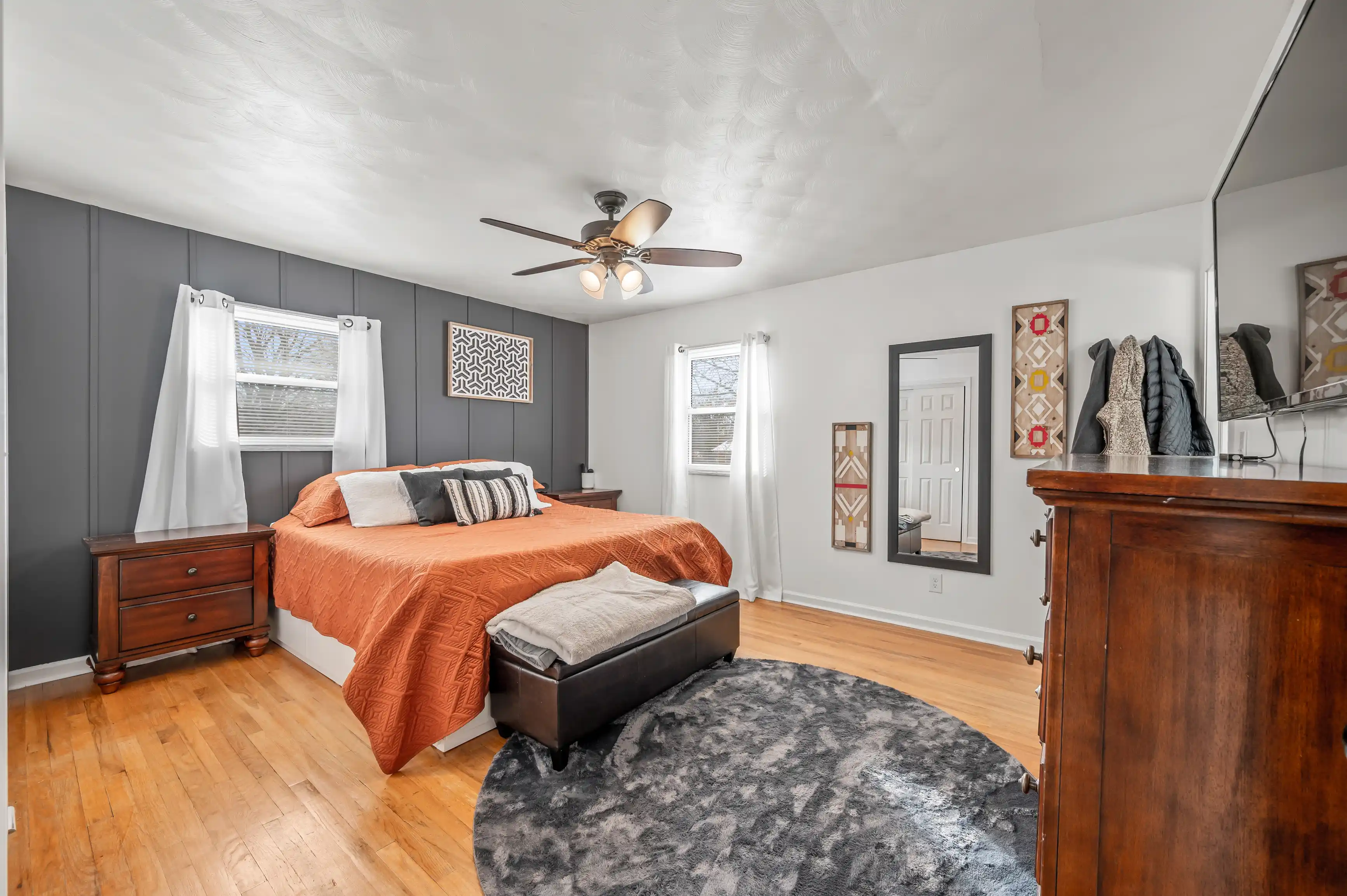 A tastefully decorated bedroom with gray walls, hardwood floors, a burnt orange bedspread, and a central ceiling fan. Various pieces of wooden furniture and decorative wall art complete the room's modern aesthetic.