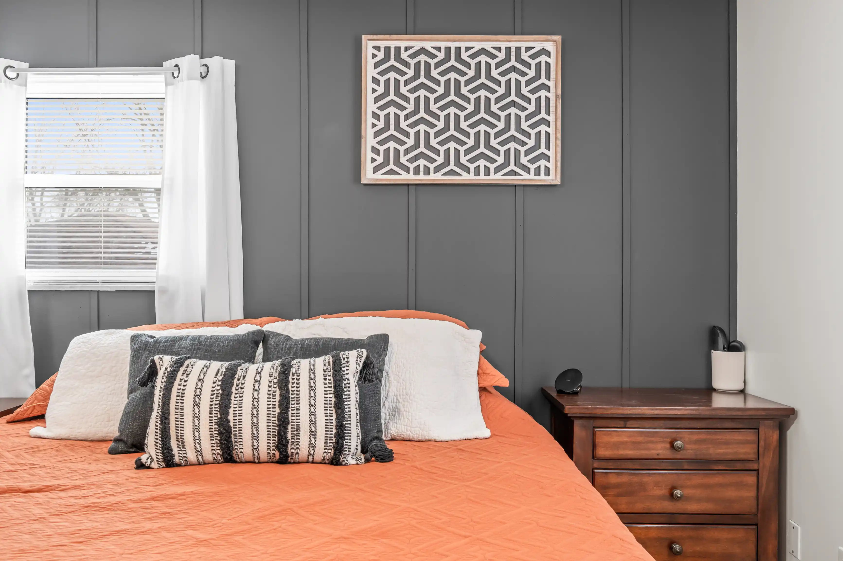 Modern bedroom with an orange bedspread, decorative pillows, a wooden nightstand, and a geometric pattern art piece on a gray wall.