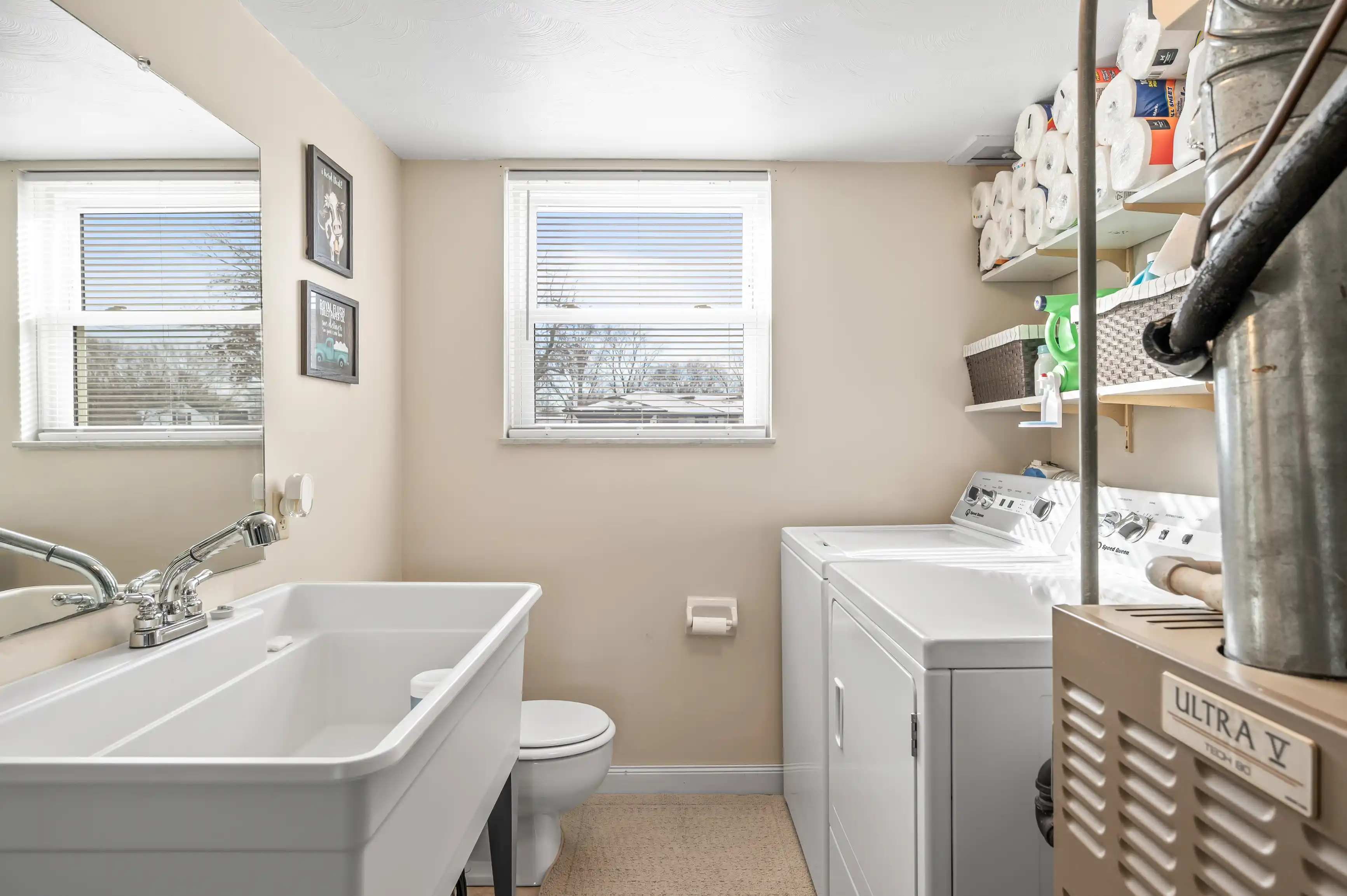 A bright laundry room with a utility sink, washing machine, dryer, and toilet, with natural light coming in through two windows.