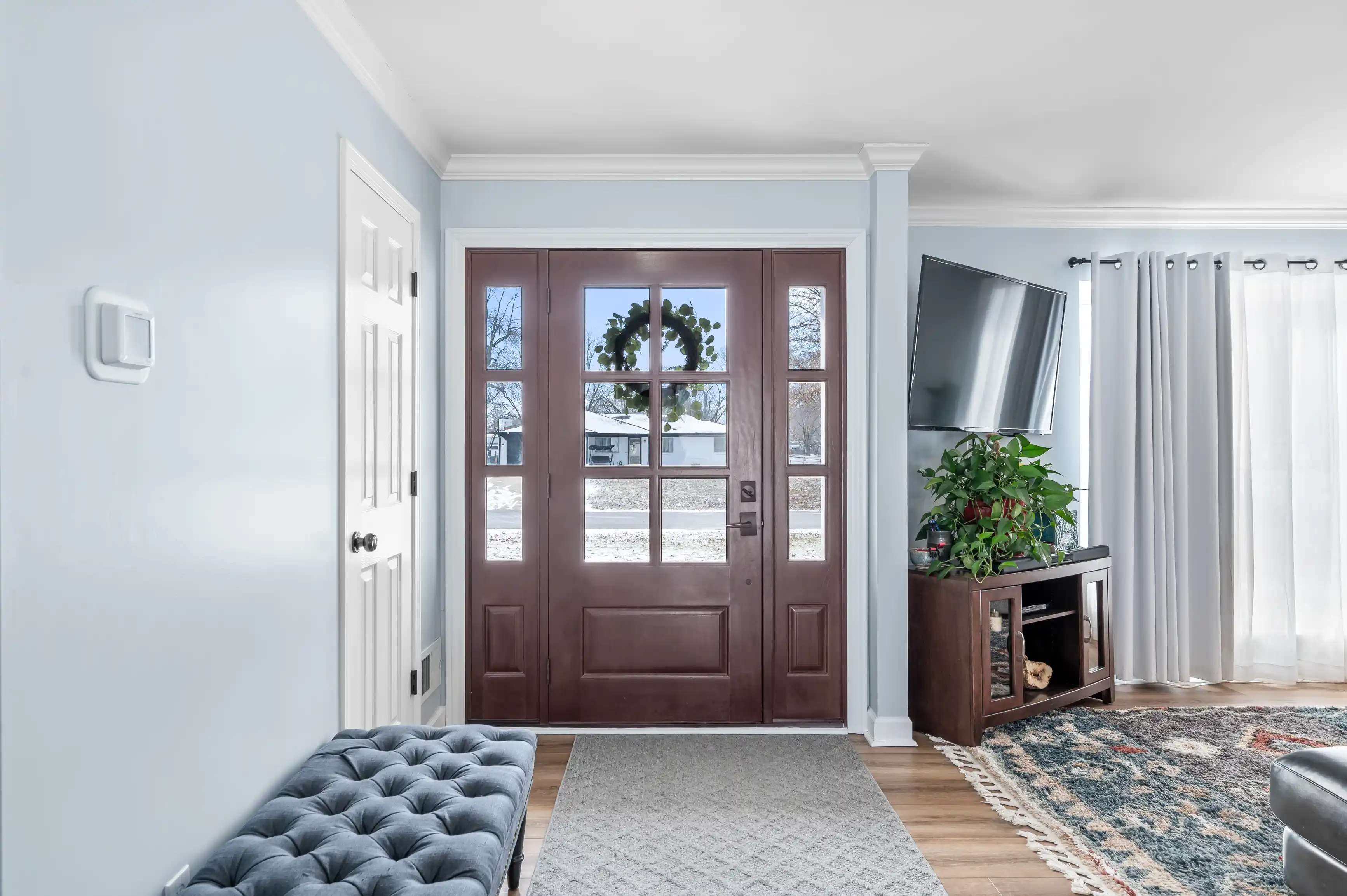 Bright entryway with a wooden front door flanked by sidelights, opening to a snow-covered exterior, with a tufted bench on one side and a television console with plants on the other.