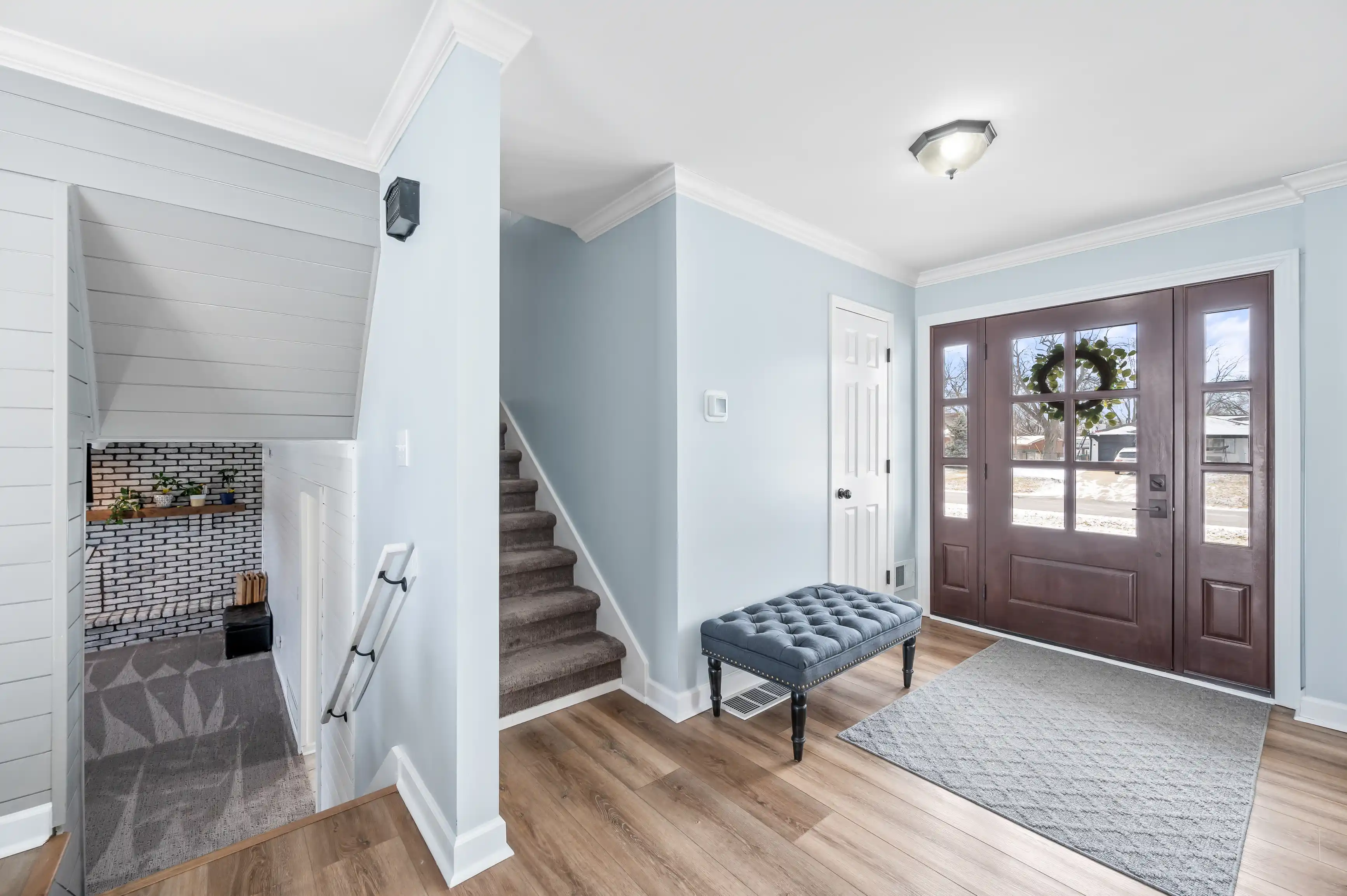 Bright and airy entryway with wooden floors, a light blue color scheme, a staircase to the left, a tiled front facing fireplace, and a large wooden front door with glass panels.