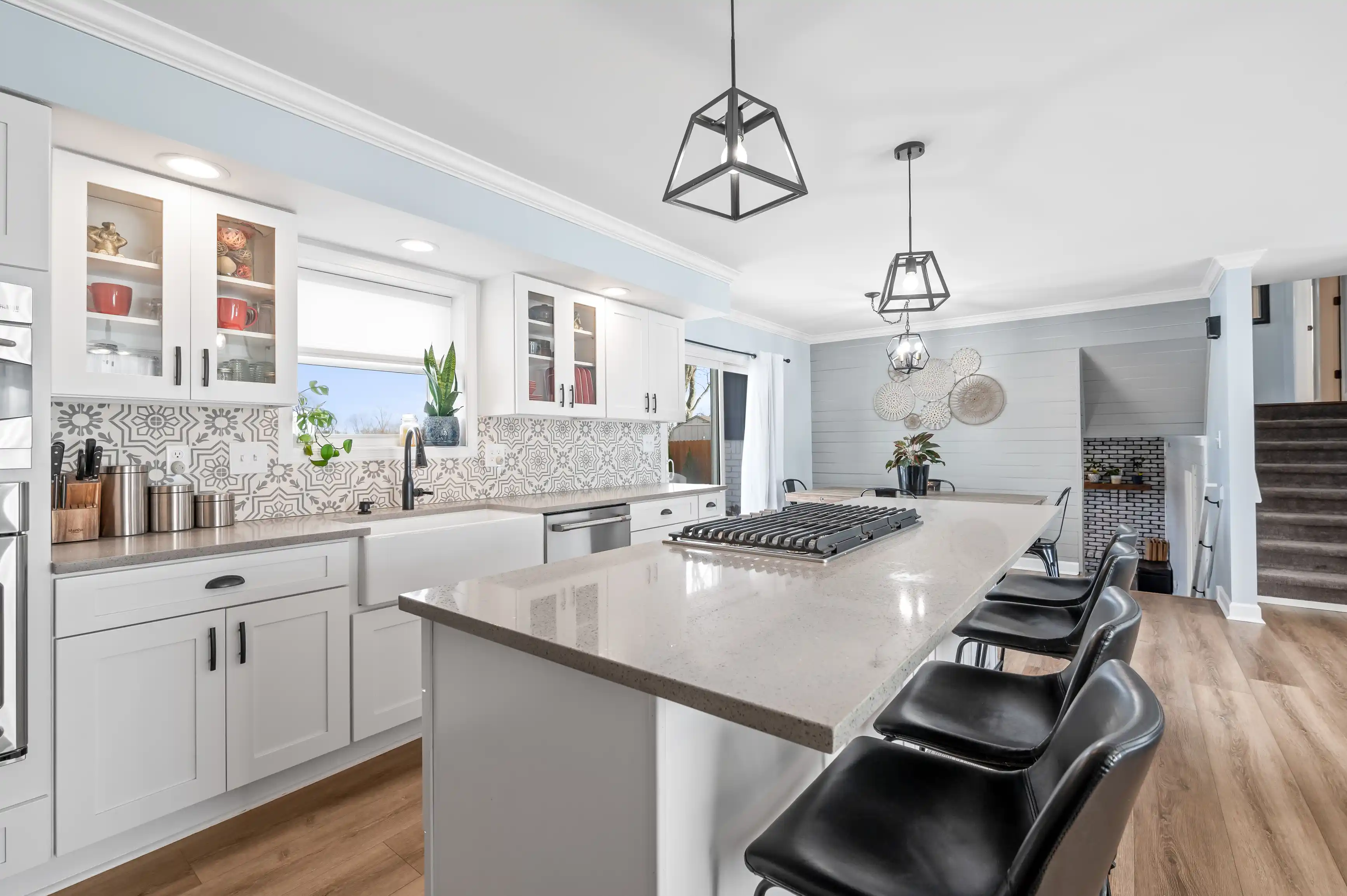 Modern kitchen interior with white cabinets, patterned backsplash, central island with black stools, pendant lights, and a view to a dining area.