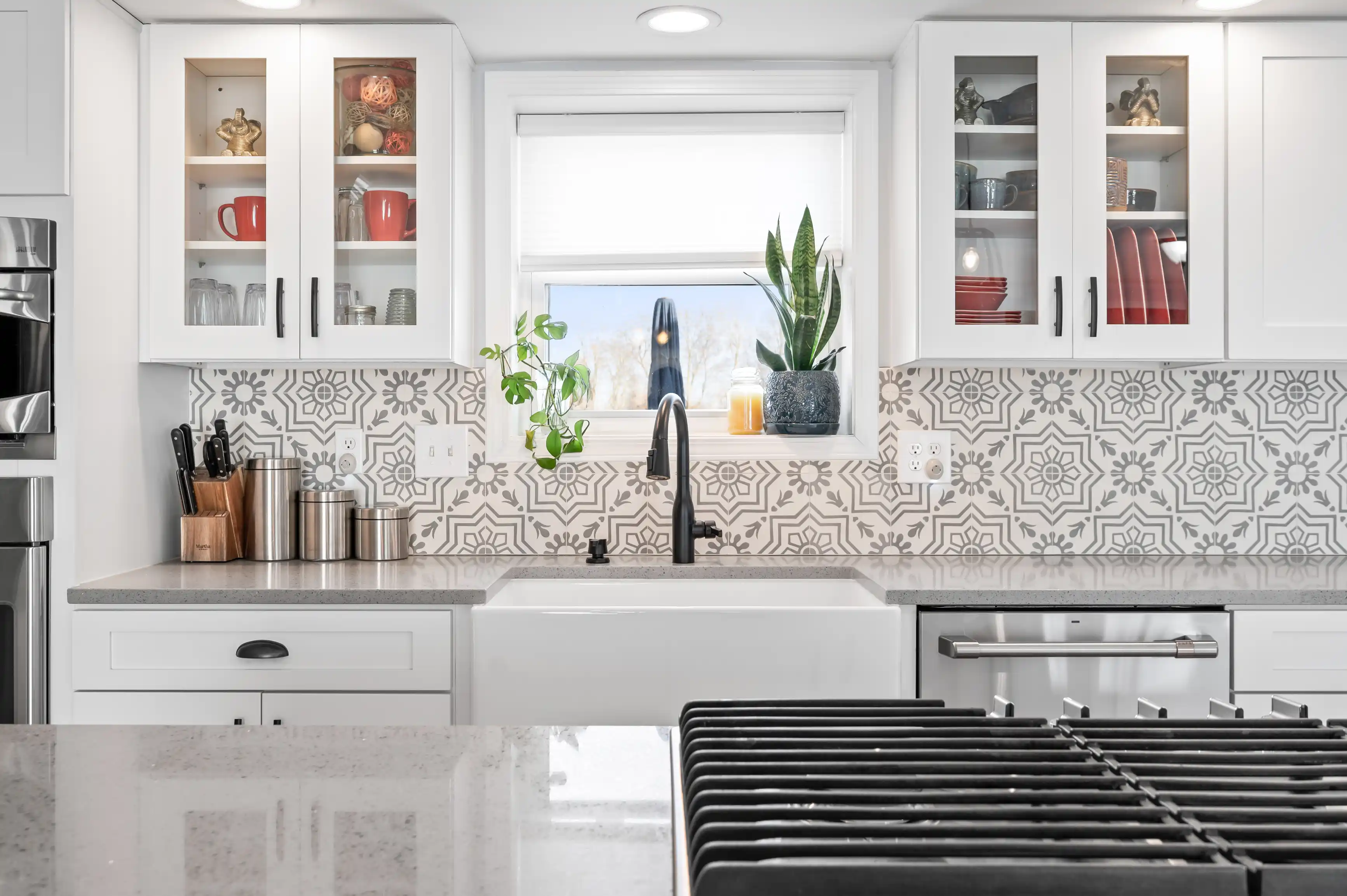 Modern kitchen interior with white cabinets, patterned backsplash, farmhouse sink, and stainless steel appliances.