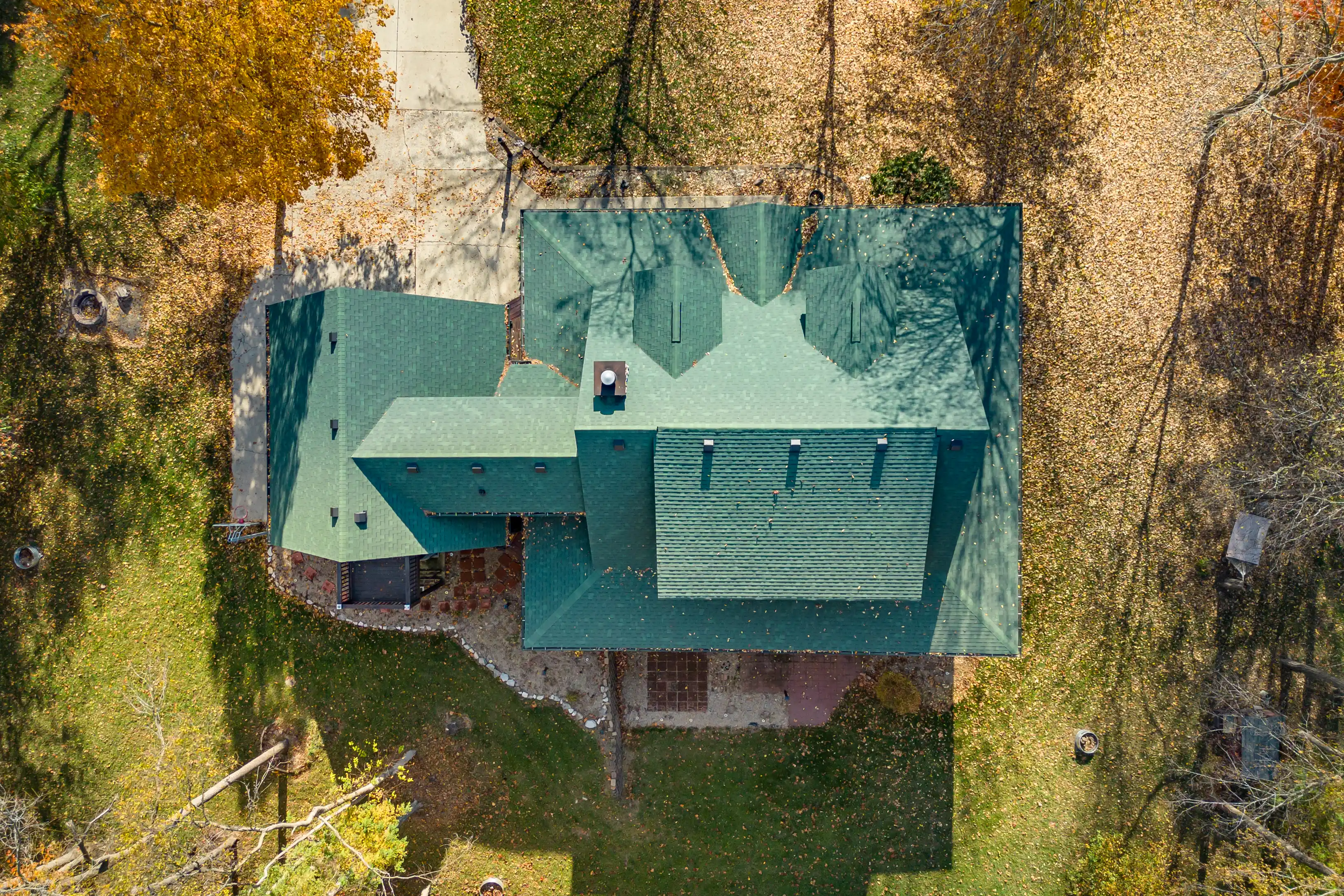 Aerial view of a house with a green roof surrounded by trees with autumn foliage and scattered leaves on the ground.