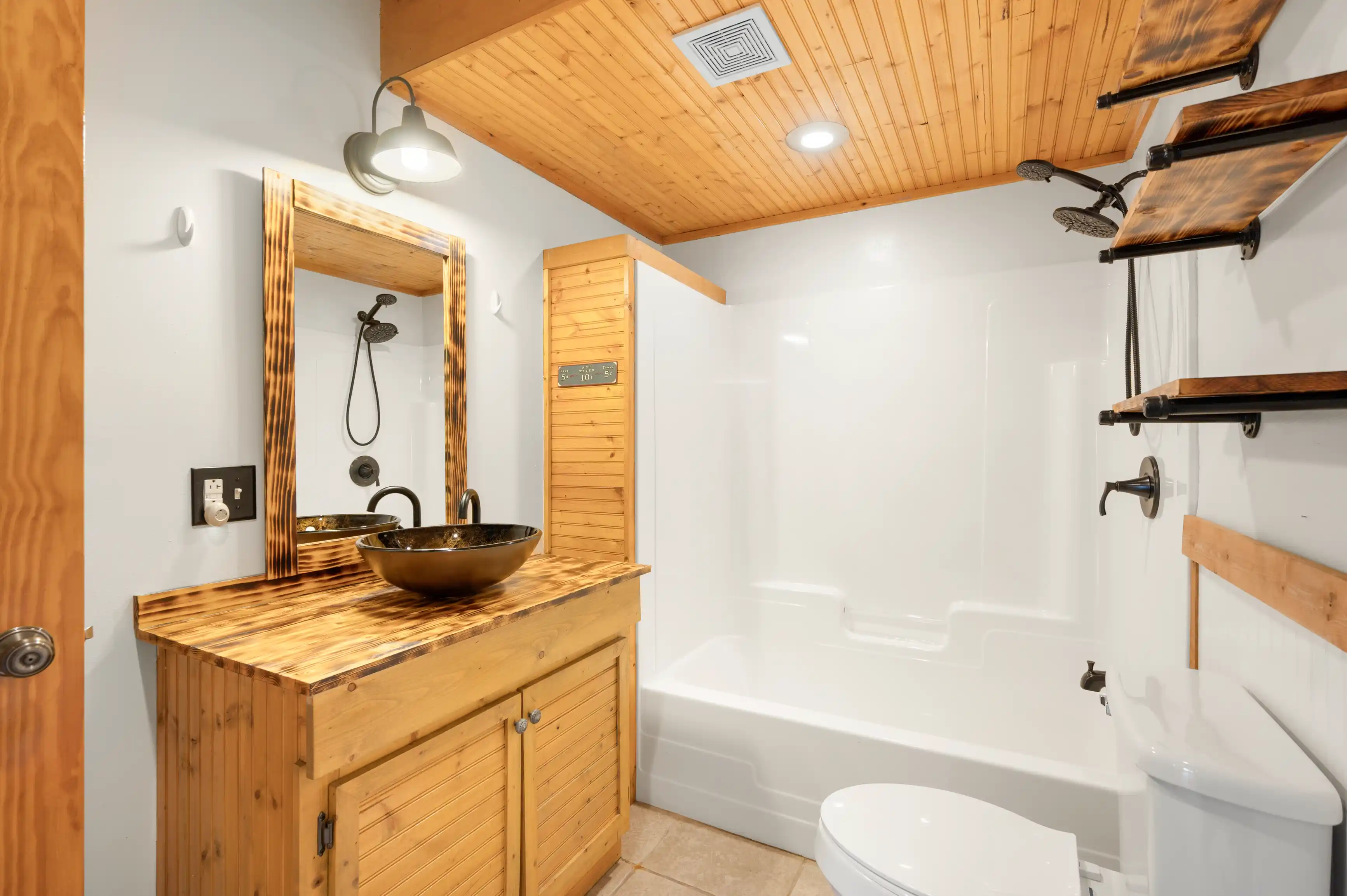 A well-lit modern bathroom with wood accents featuring a bowl sink on a wooden vanity, a large mirror, a bathtub with shower, a toilet, and a wooden sauna cabin.