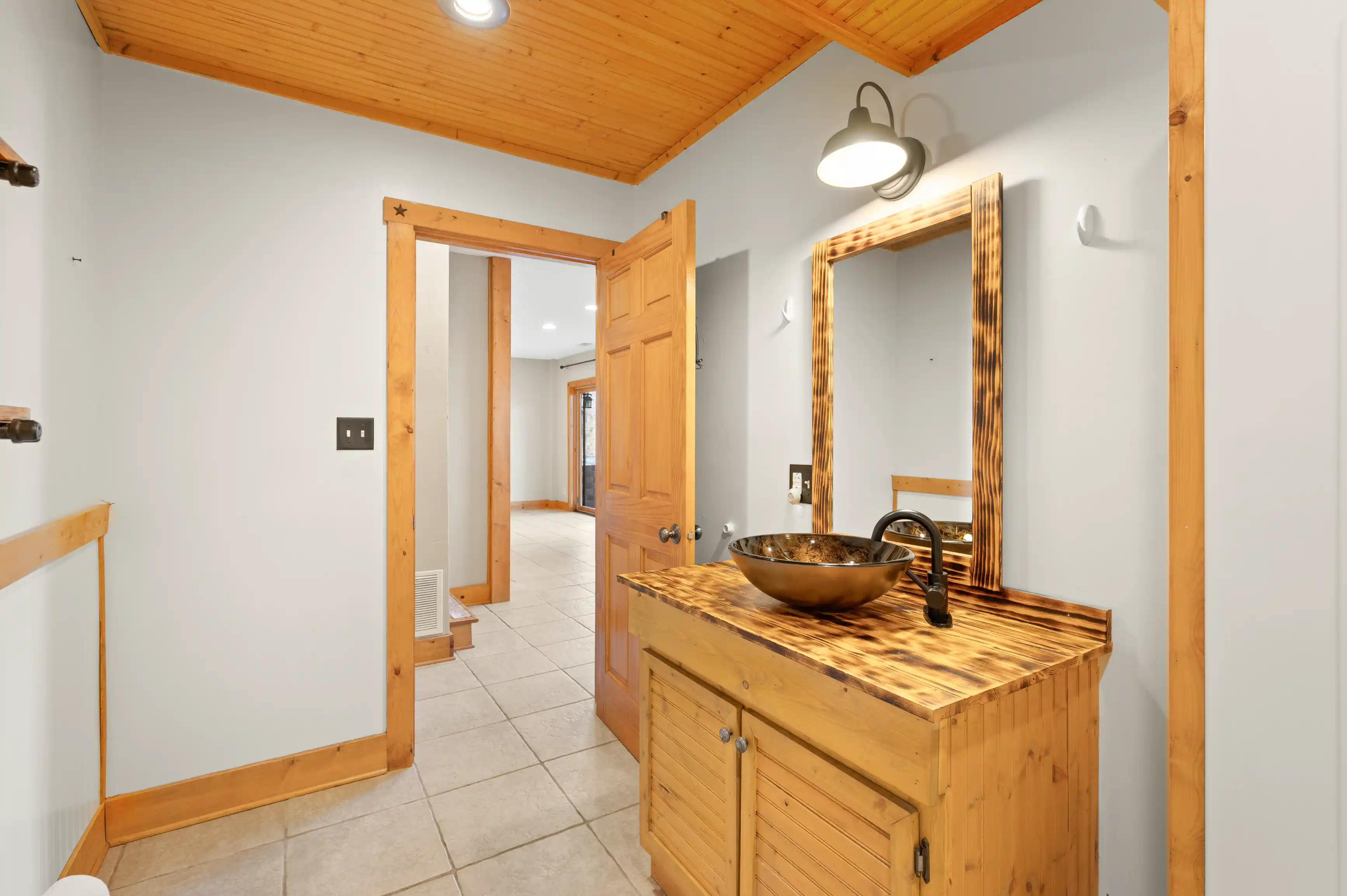 Interior view of a hallway with wooden ceiling and trim, featuring a vanity with a vessel sink and mirror, leading to another room with open doors.