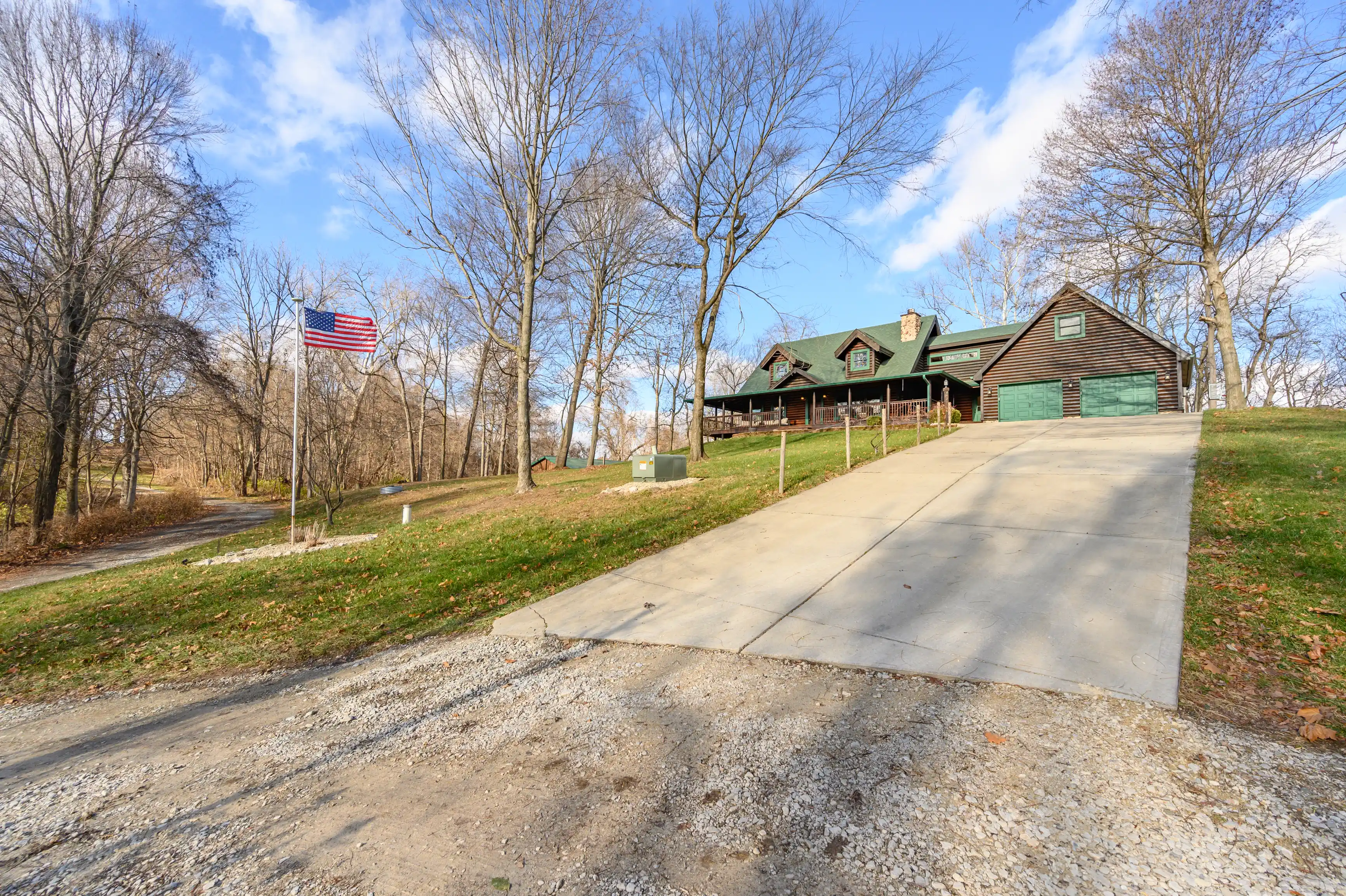 A rustic log cabin with a green roof and attached garage located in a leafless wooded area with an American flag on a flagpole, under a blue sky with scattered clouds.