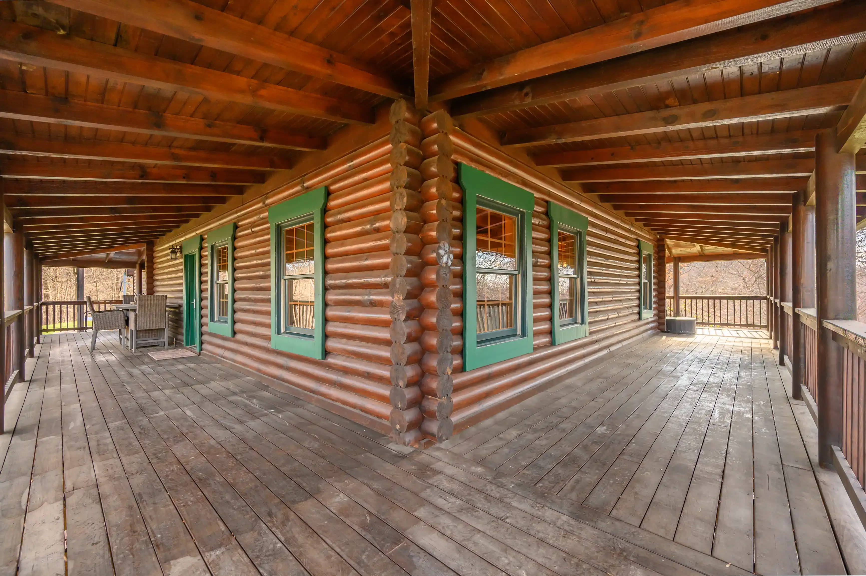 Spacious wooden porch of a log cabin with green-trimmed windows and outdoor furniture overlooking a forested area.
