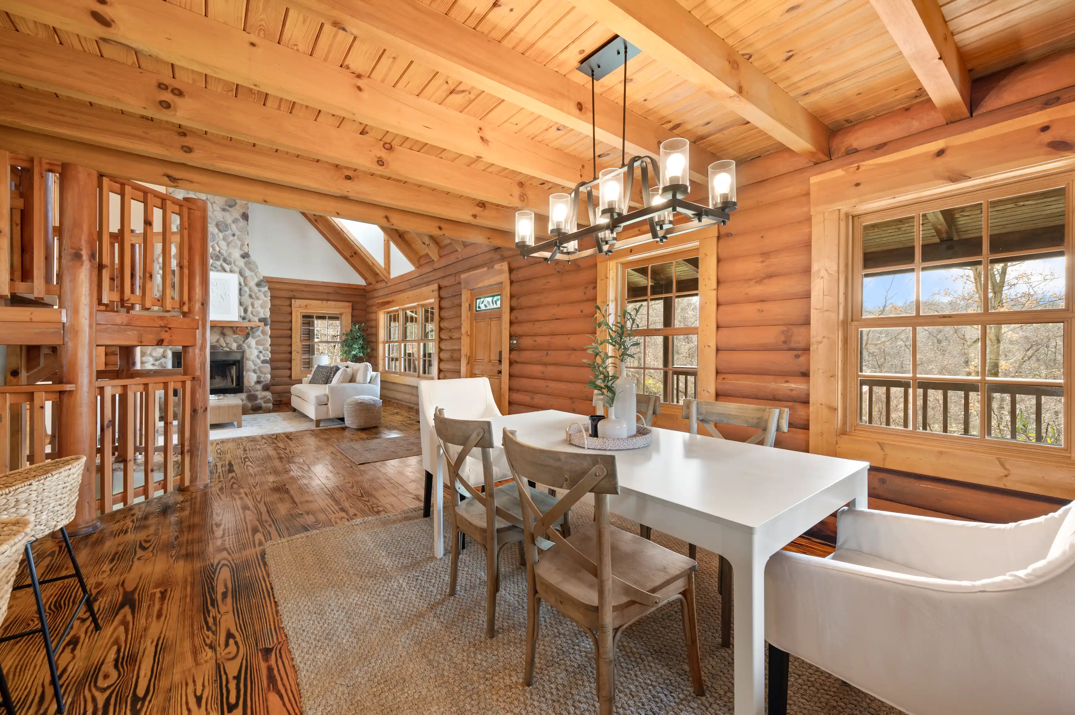 Cozy log cabin interior with exposed wooden beams, a dining area with a white table and chairs, and a living room with a stone fireplace in the background, natural light streaming through large windows.