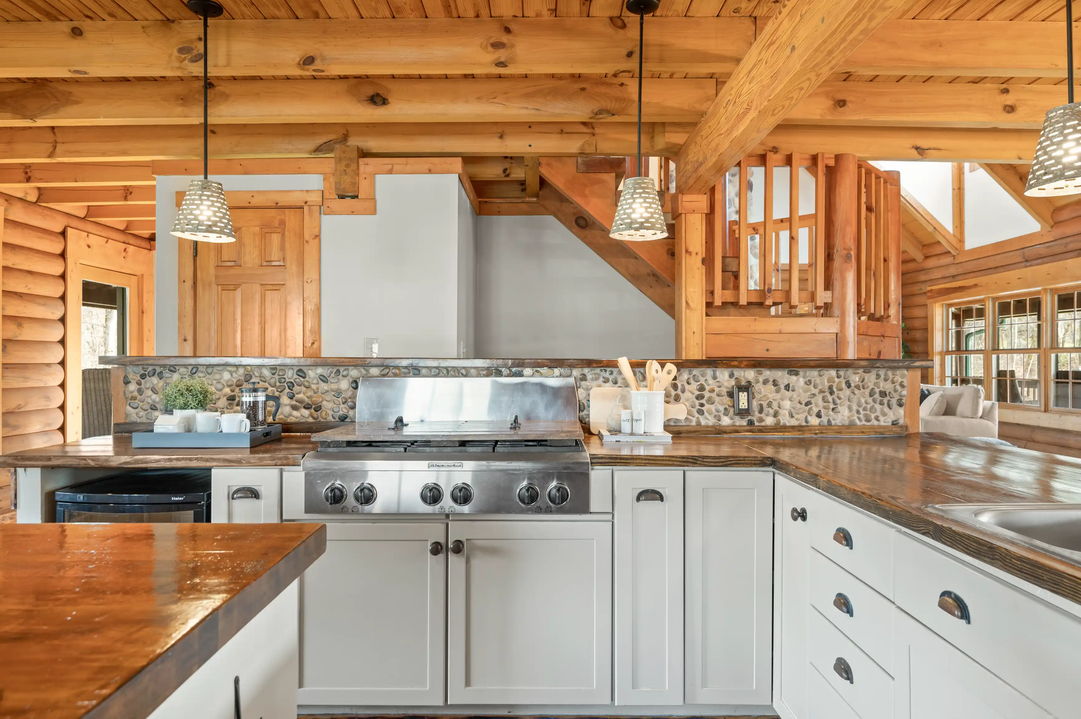 Rustic wooden cabin kitchen interior with log construction, stone backsplash, stainless steel stove, wooden countertops, and hanging pendant lights.