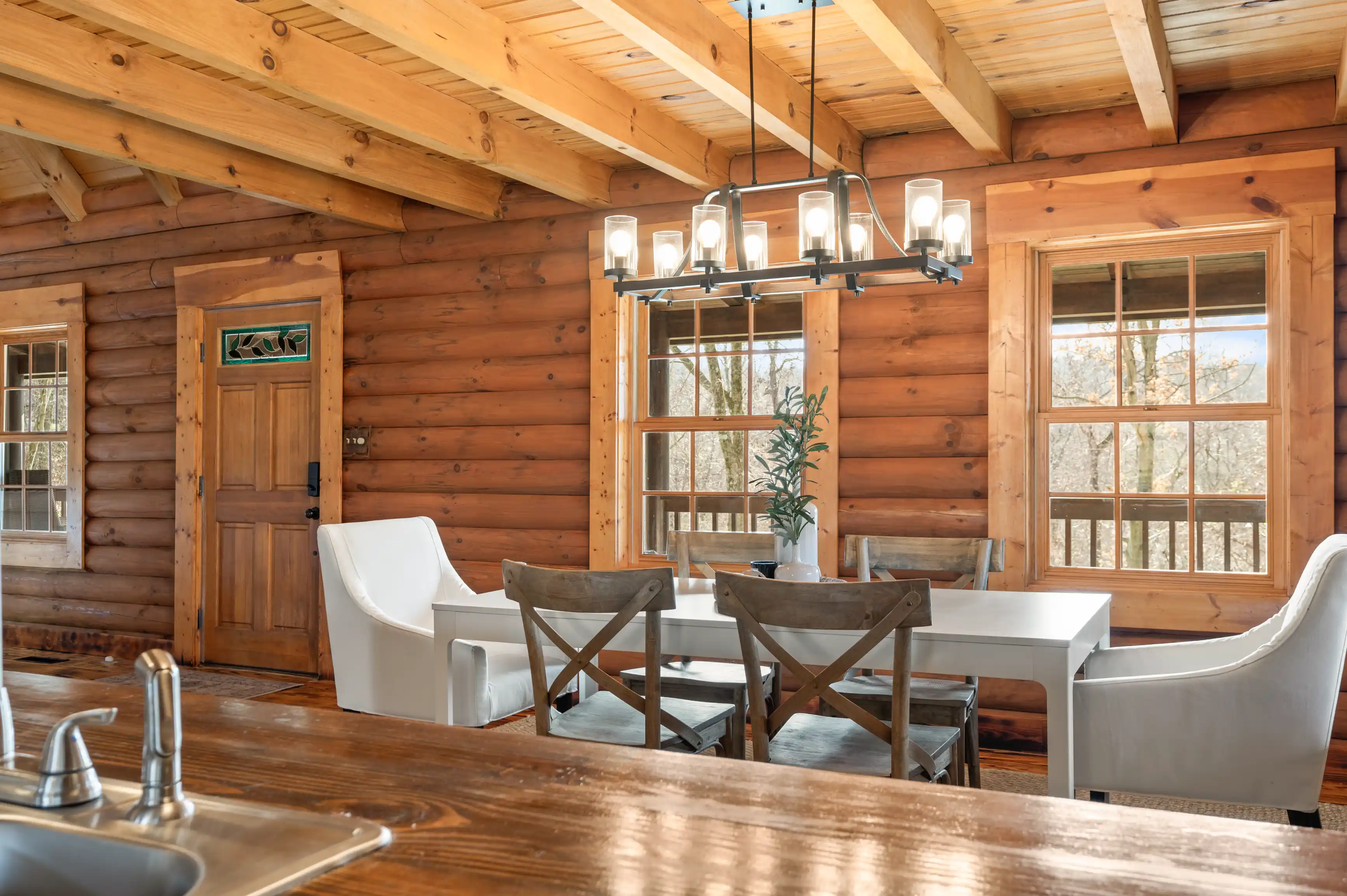 Rustic cabin interior with log walls, exposed wooden beams, and a dining area with modern chairs under a contemporary chandelier.