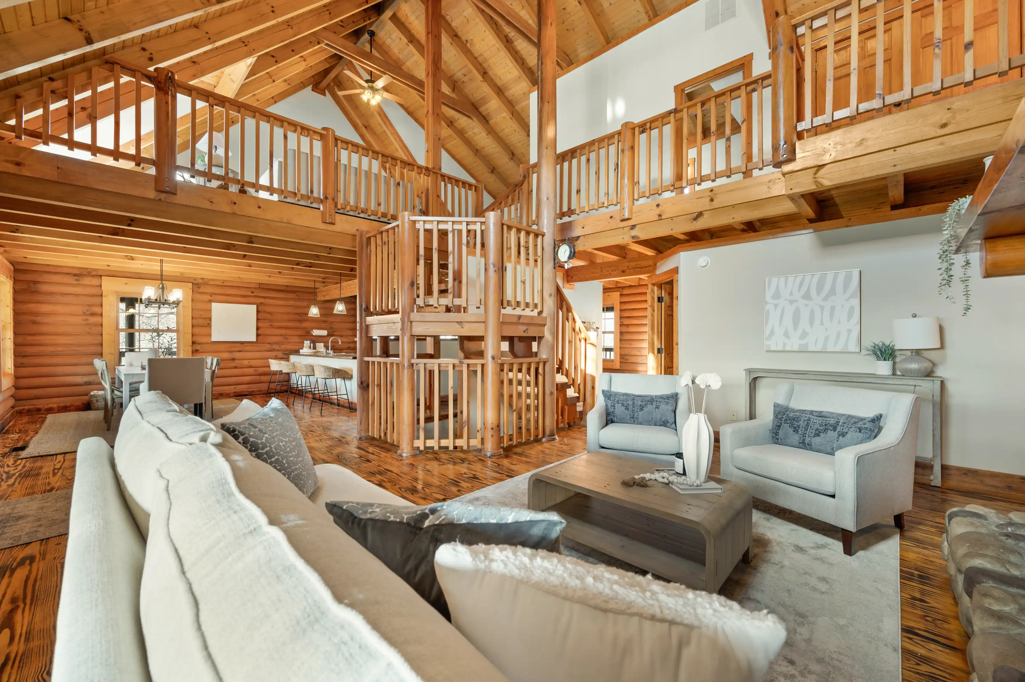 Spacious log cabin interior with a high vaulted ceiling, exposed wooden beams, and a loft area, featuring a cozy living room setup with sofas and a dining space in the background.