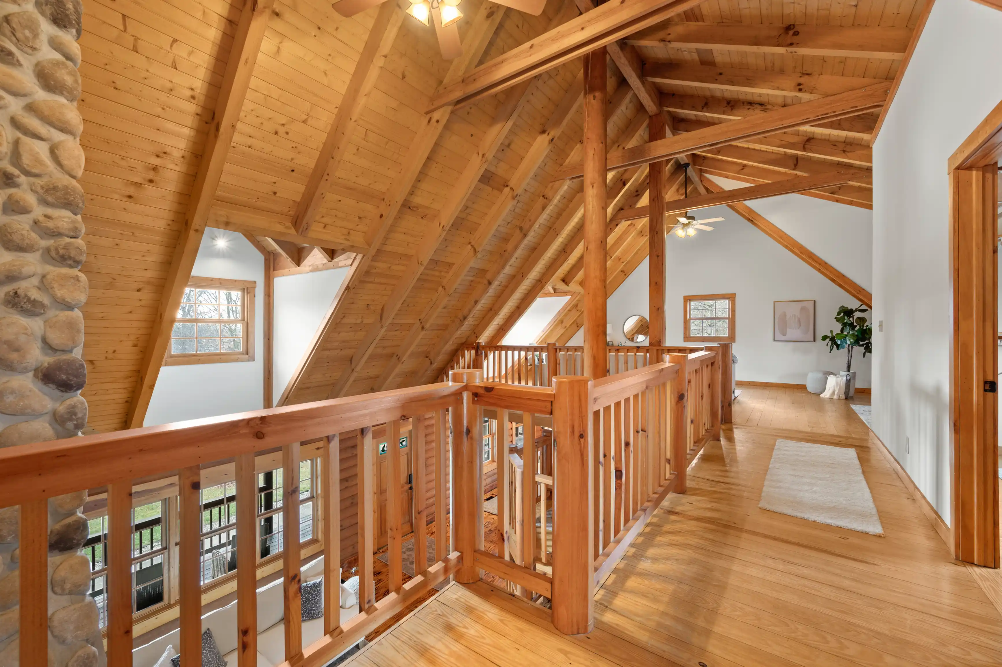 A spacious interior of a wooden attic room with exposed beams, stone column, wooden floor and guardrails, and a window letting in natural light.