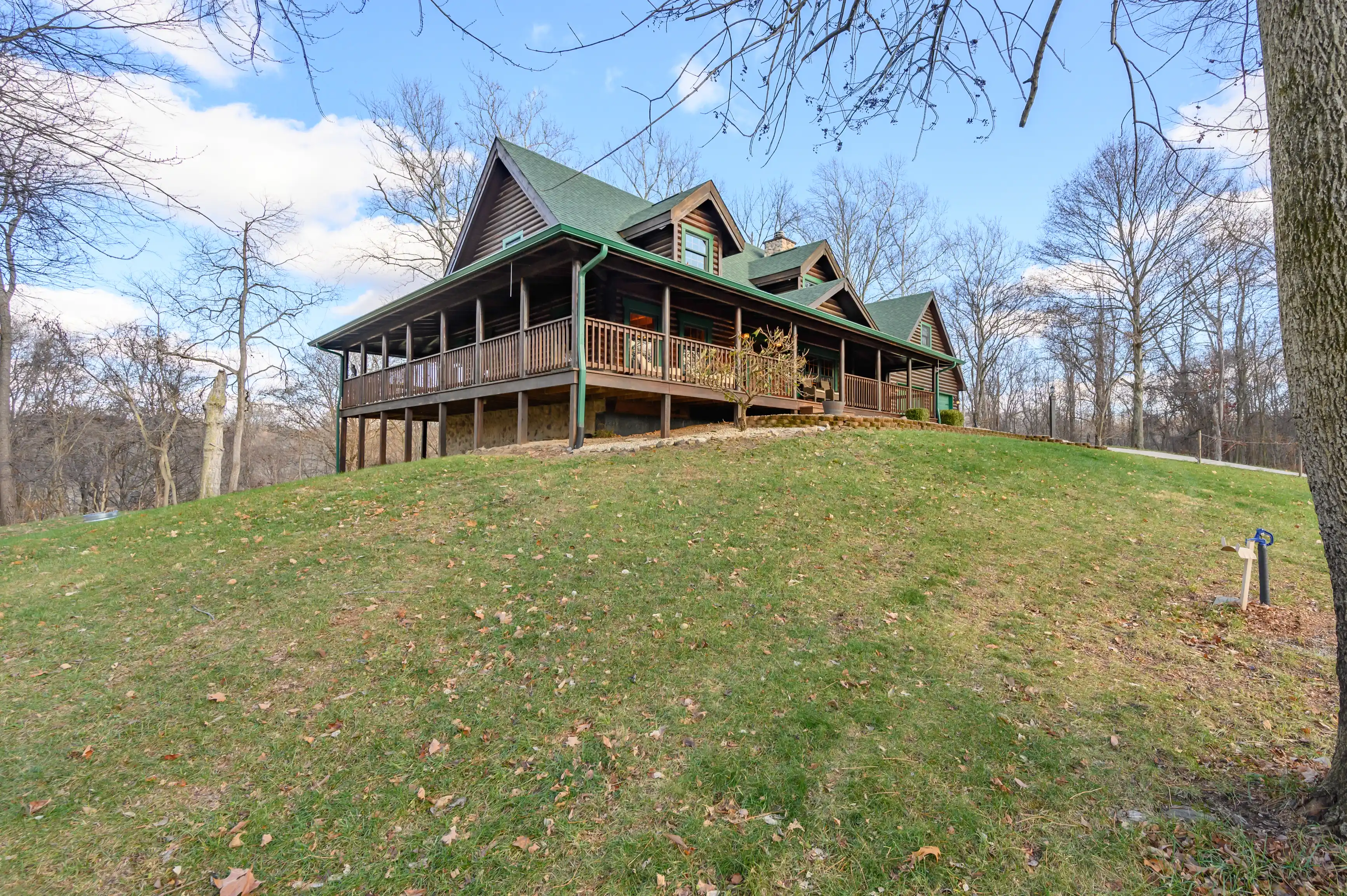 A large cabin with a green roof and an expansive wooden deck sitting on a grassy hill with bare trees in the background and a clear sky.