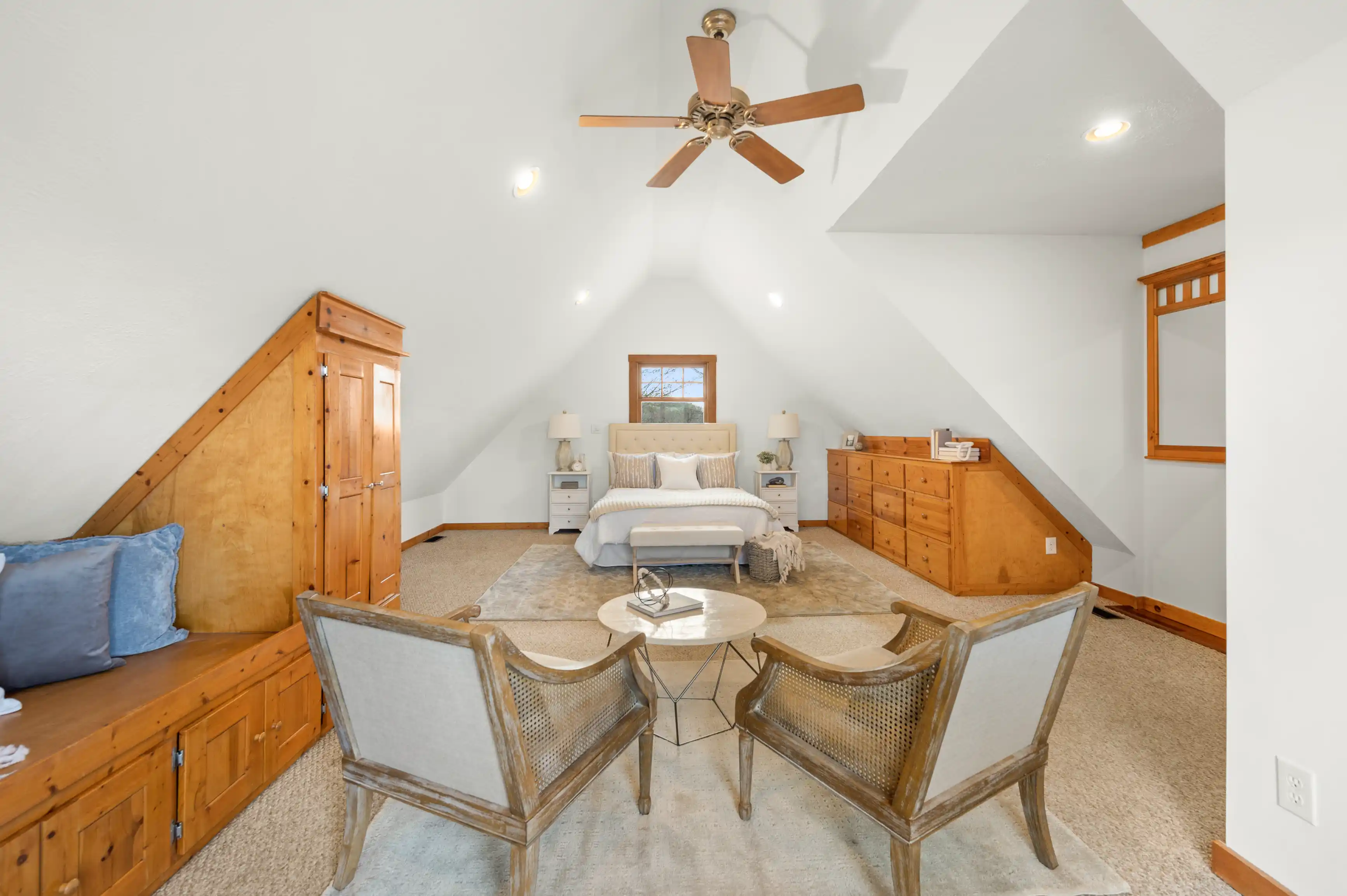 Bright and airy attic bedroom with vaulted ceilings, ceiling fan, wooden furniture including a bed with white bedding, dresser, and built-in storage bench, accented with a small round table and two chairs on a beige carpet.