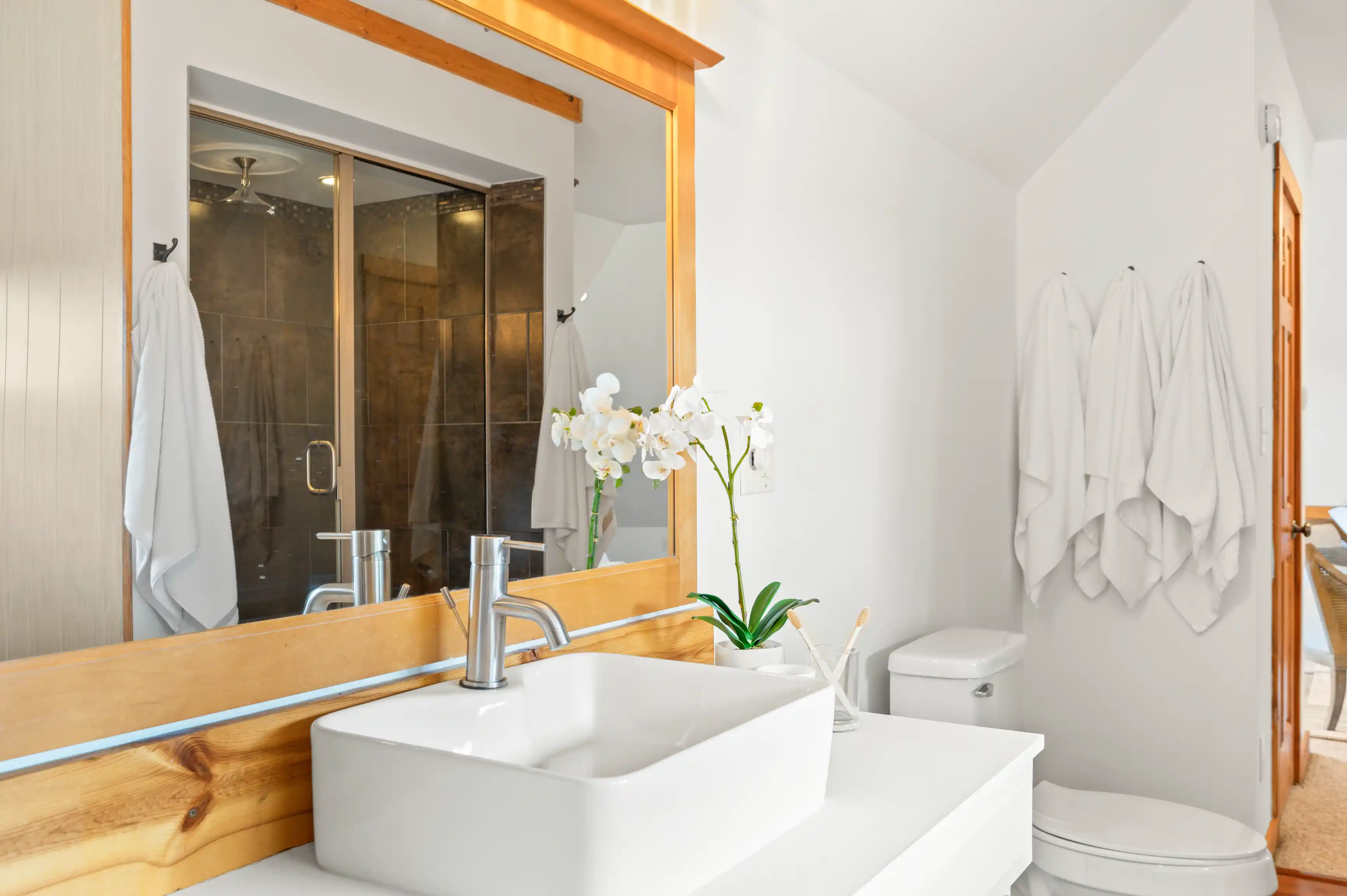 Bright modern bathroom interior with wooden countertop, rectangular vessel sink, orchid plant, shower stall with glass doors, and white towels hanging on wall hooks.
