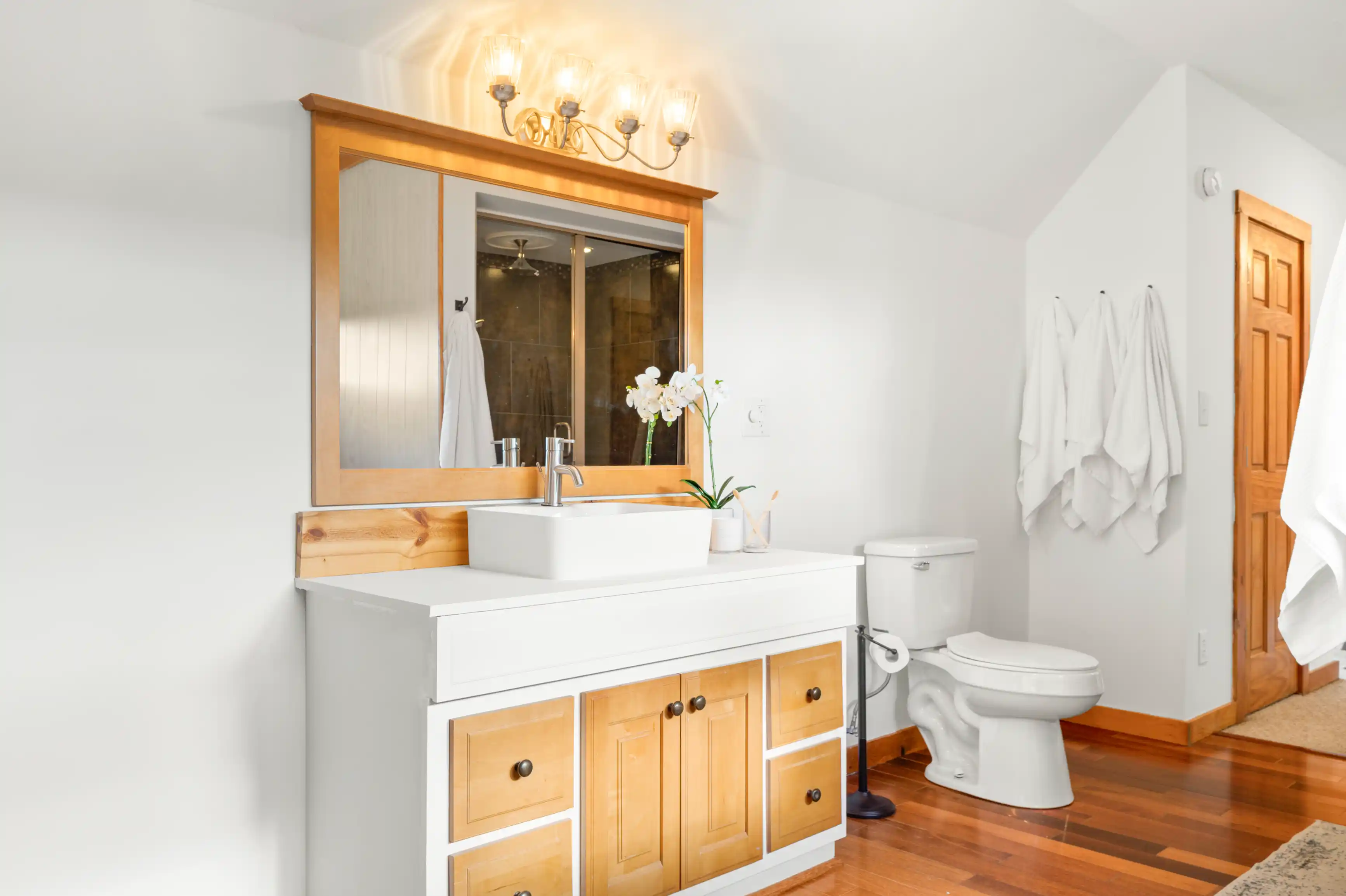 Bright and modern bathroom interior with wooden vanity cabinet, white sink, large mirror, toilet, and towels hanging on the wall.