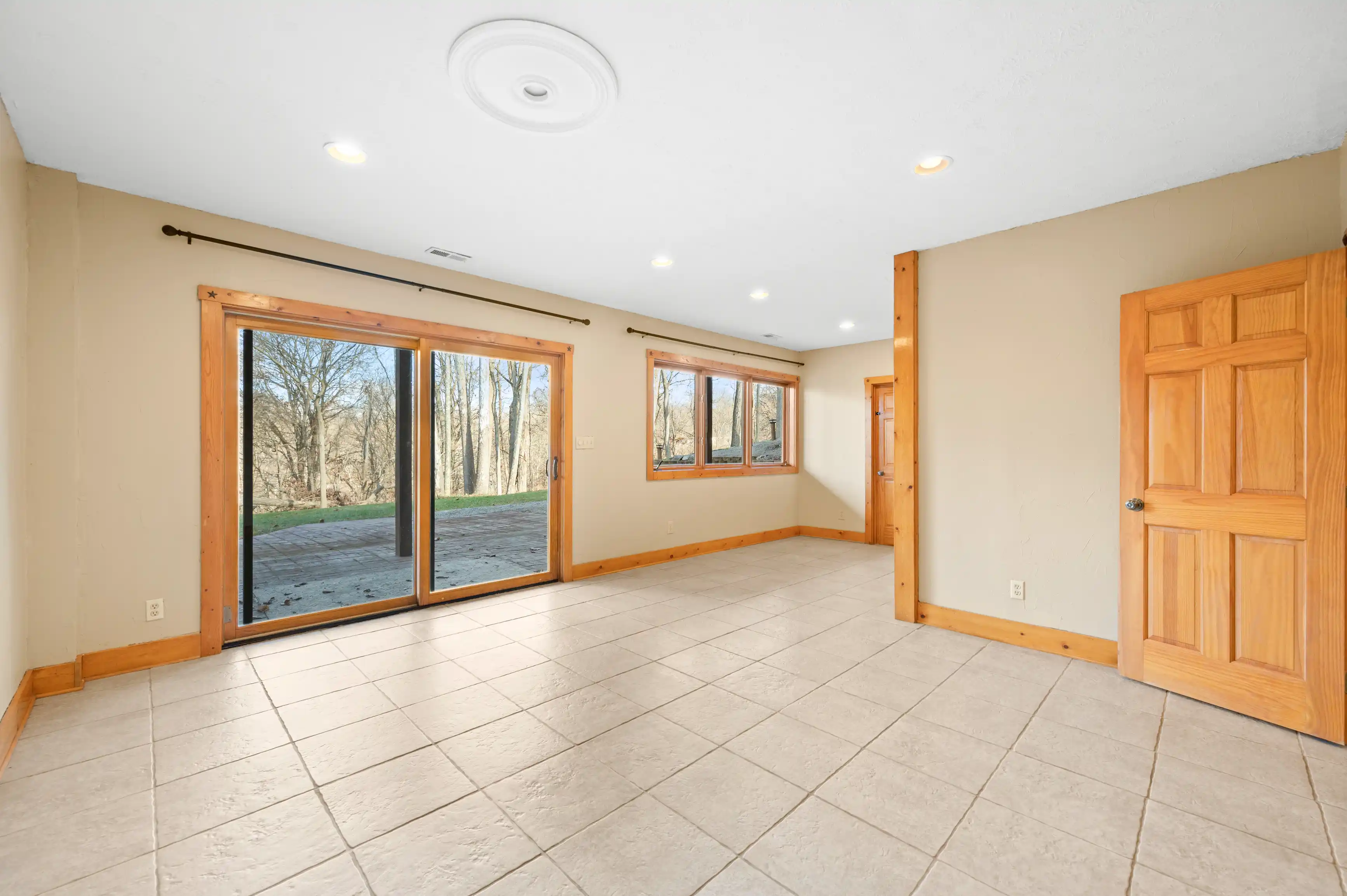 Empty room with tiled floor, wooden trim, sliding glass door to the backyard, and wooden entry door on the right.