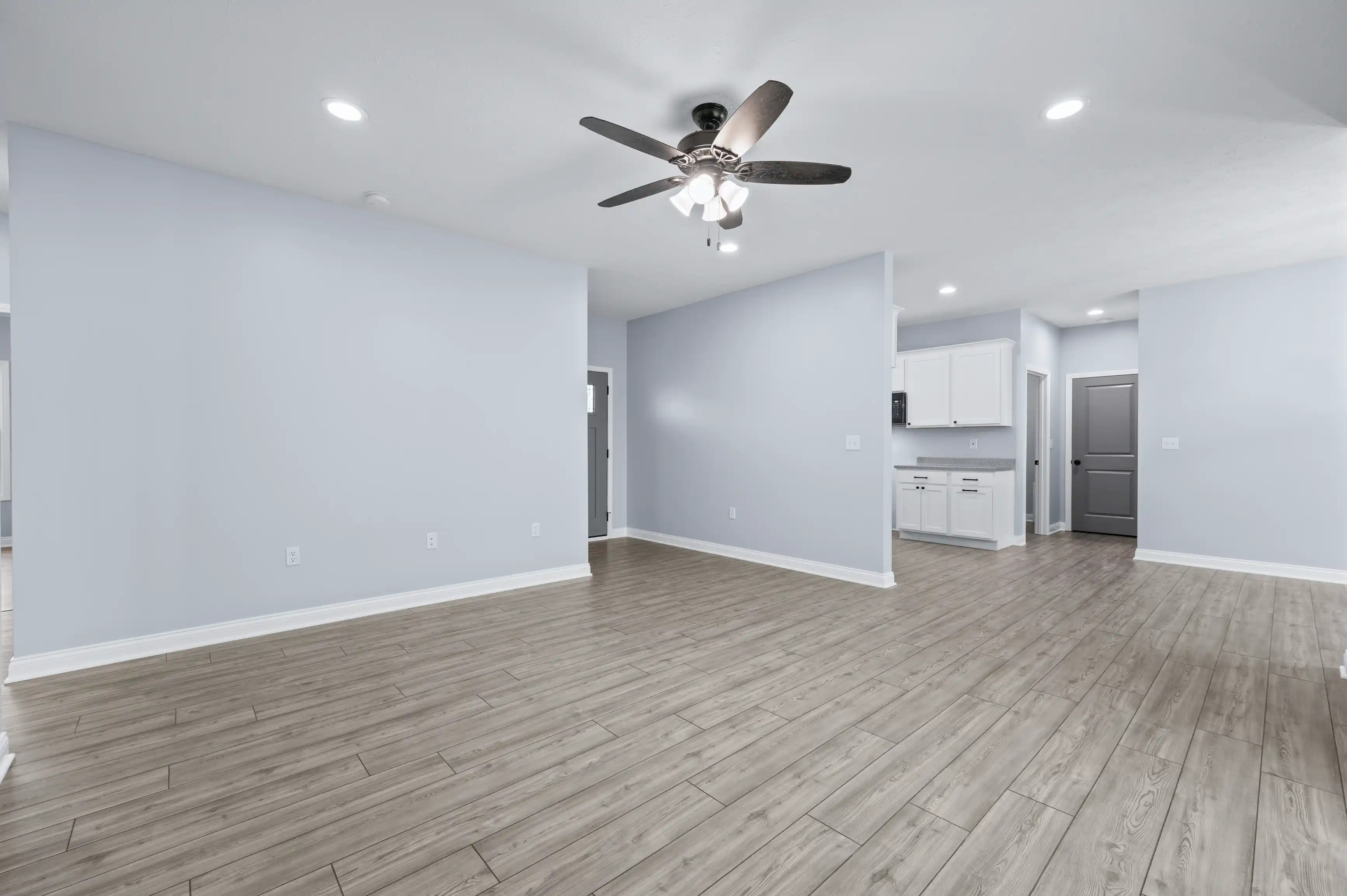 Spacious, empty room with light gray walls, dark wood flooring, white trim, a ceiling fan with lights, leading into a kitchen area with white cabinets and appliances.