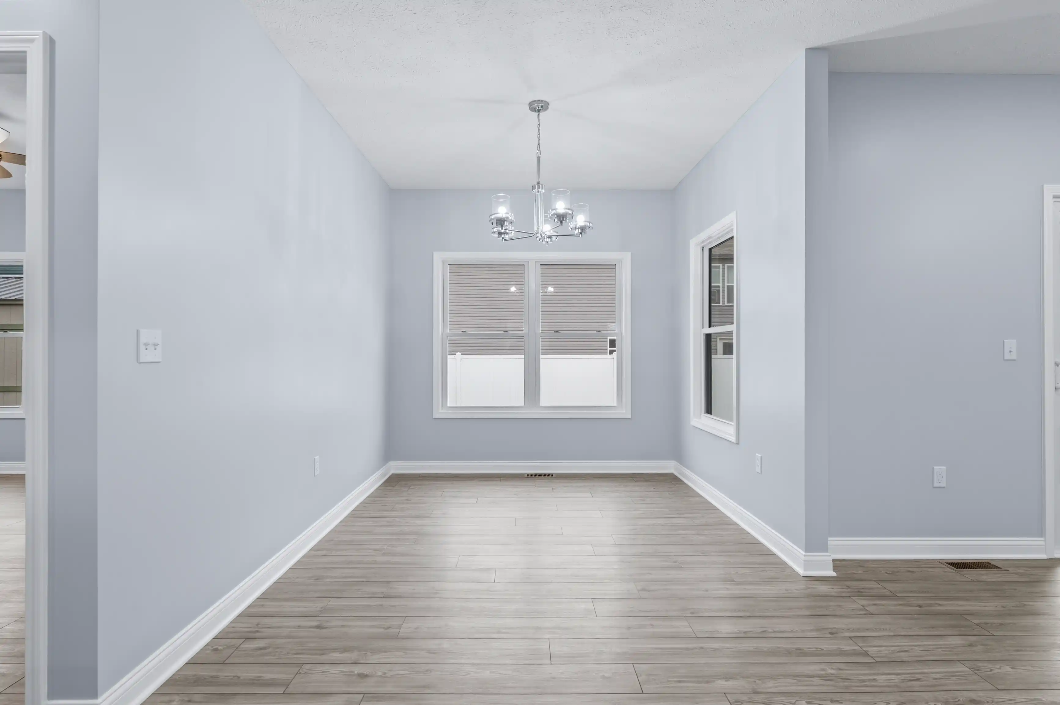 Empty room with light blue walls, a window with blinds, a silver chandelier, and laminate wood flooring.