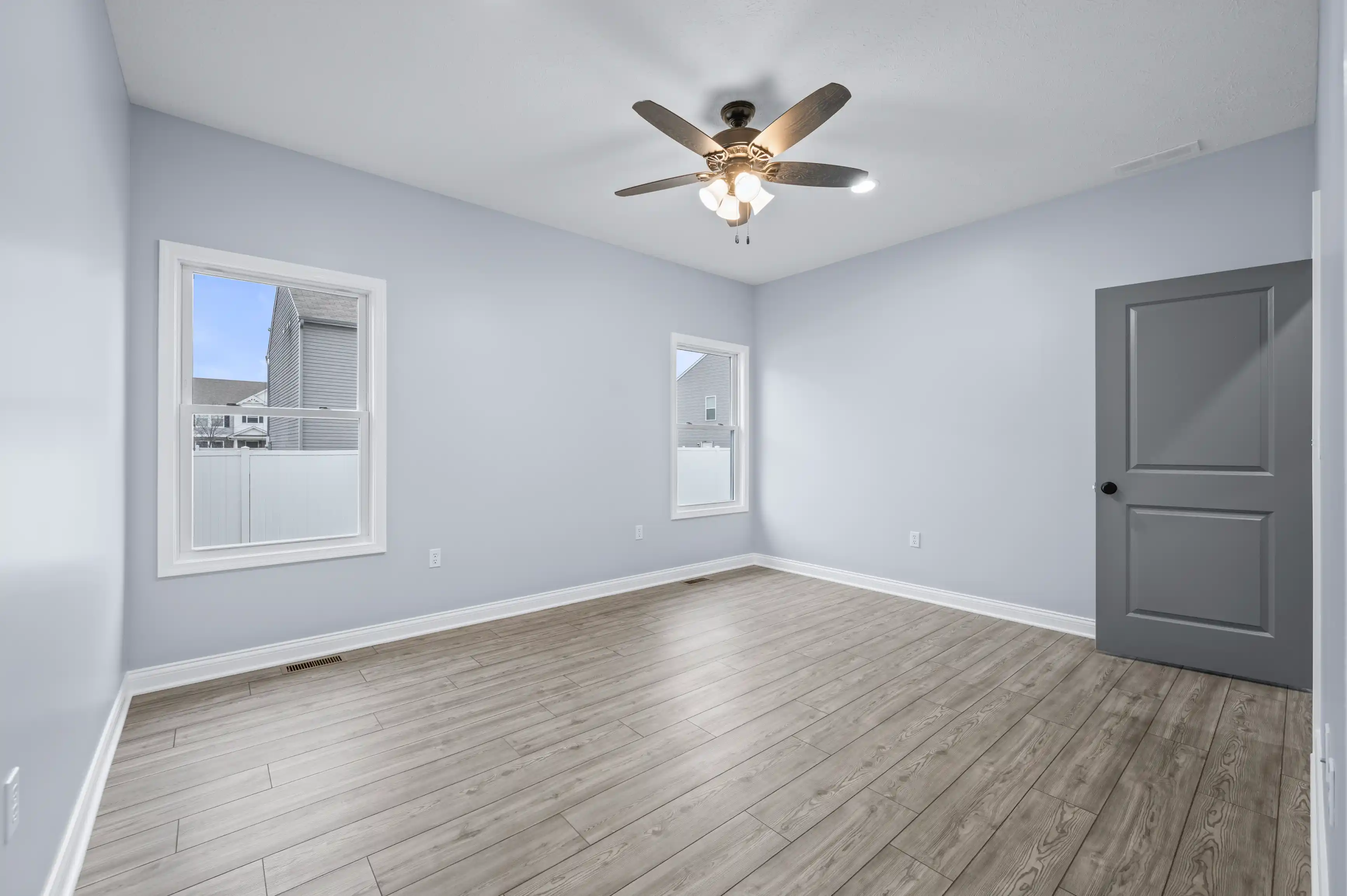 Spacious empty room with light gray walls, hardwood floors, two windows, and a ceiling fan with lights.