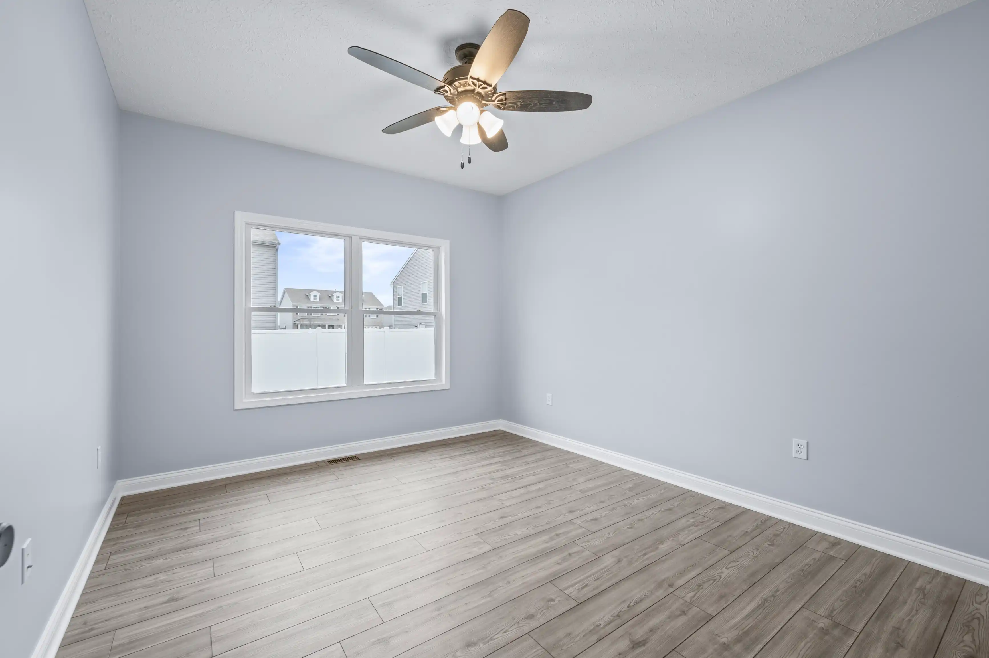 Empty room with light grey walls, ceiling fan with lights, and wood laminate flooring, featuring a large window with a view of neighboring houses.