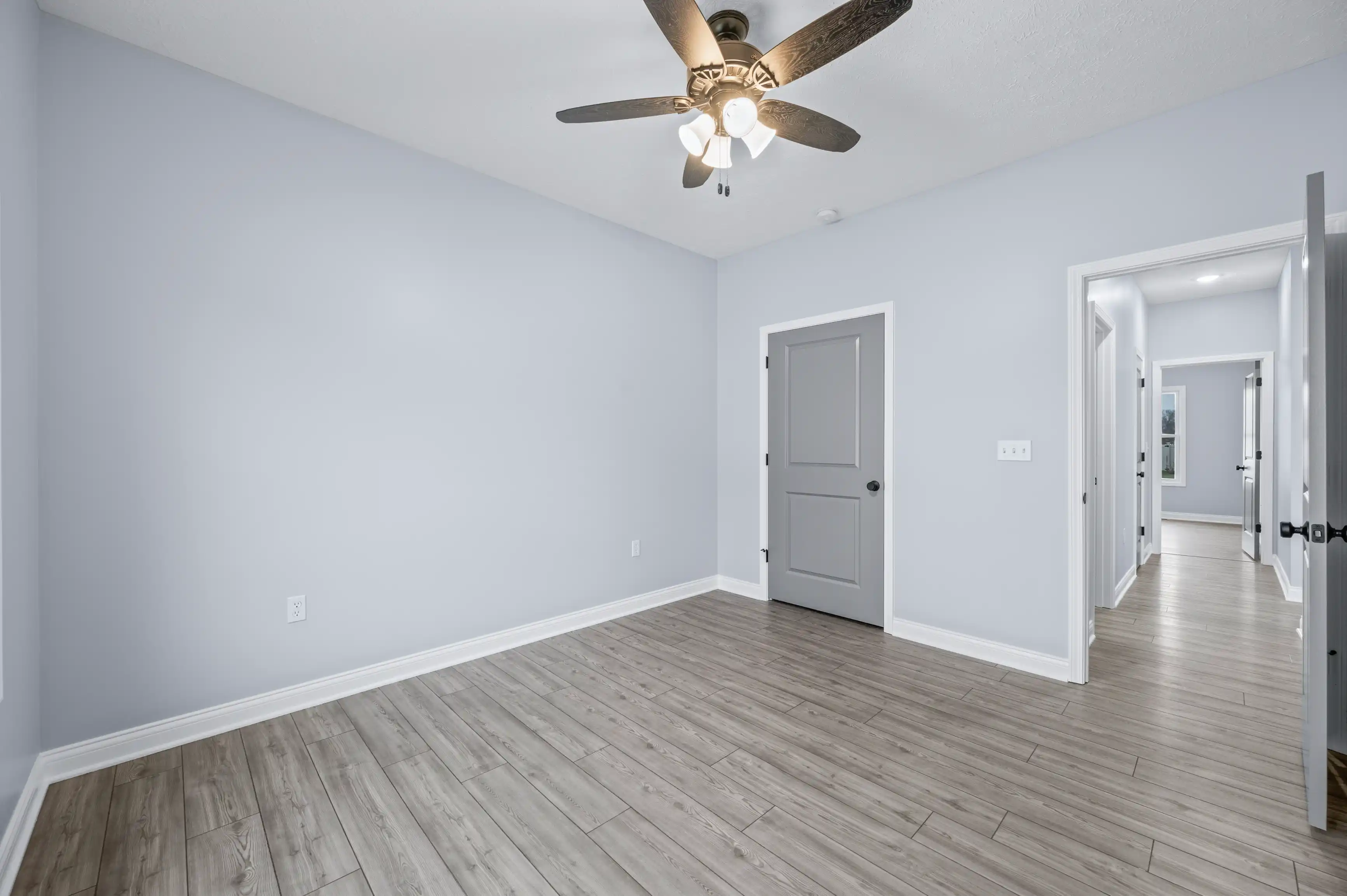 Spacious empty room with pale blue walls, wooden laminate flooring, a ceiling fan with lights, and multiple open doors leading to other parts of the house.