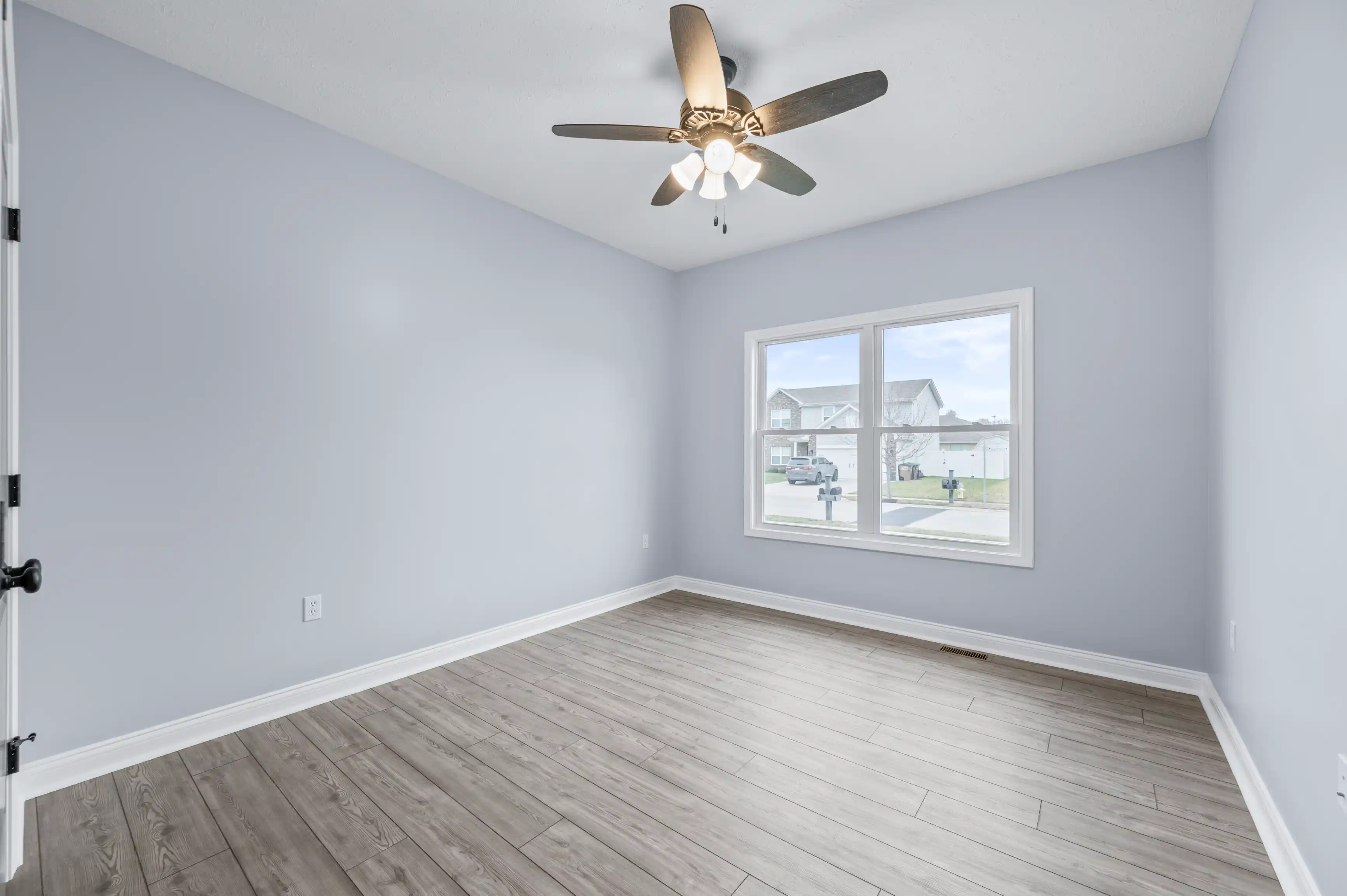 Empty room with light gray walls, wood flooring, and a ceiling fan, featuring a large window with a view of the neighborhood.