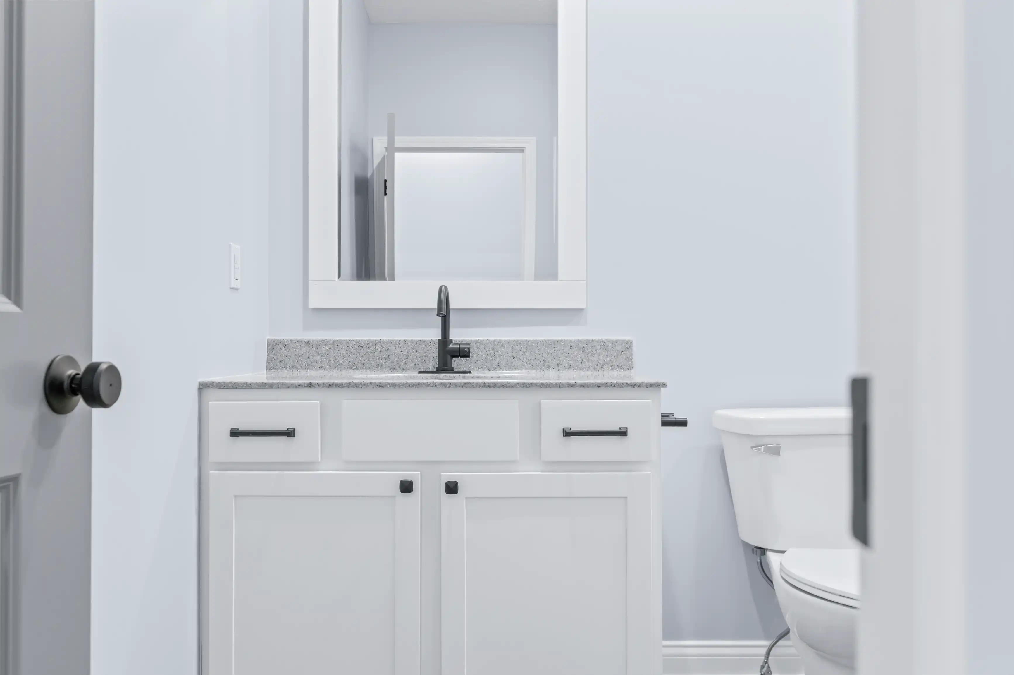 A modern, clean bathroom interior with a white vanity cabinet featuring a granite countertop and sink, a rectangular mirror above, and a toilet to the right, all against light blue walls.