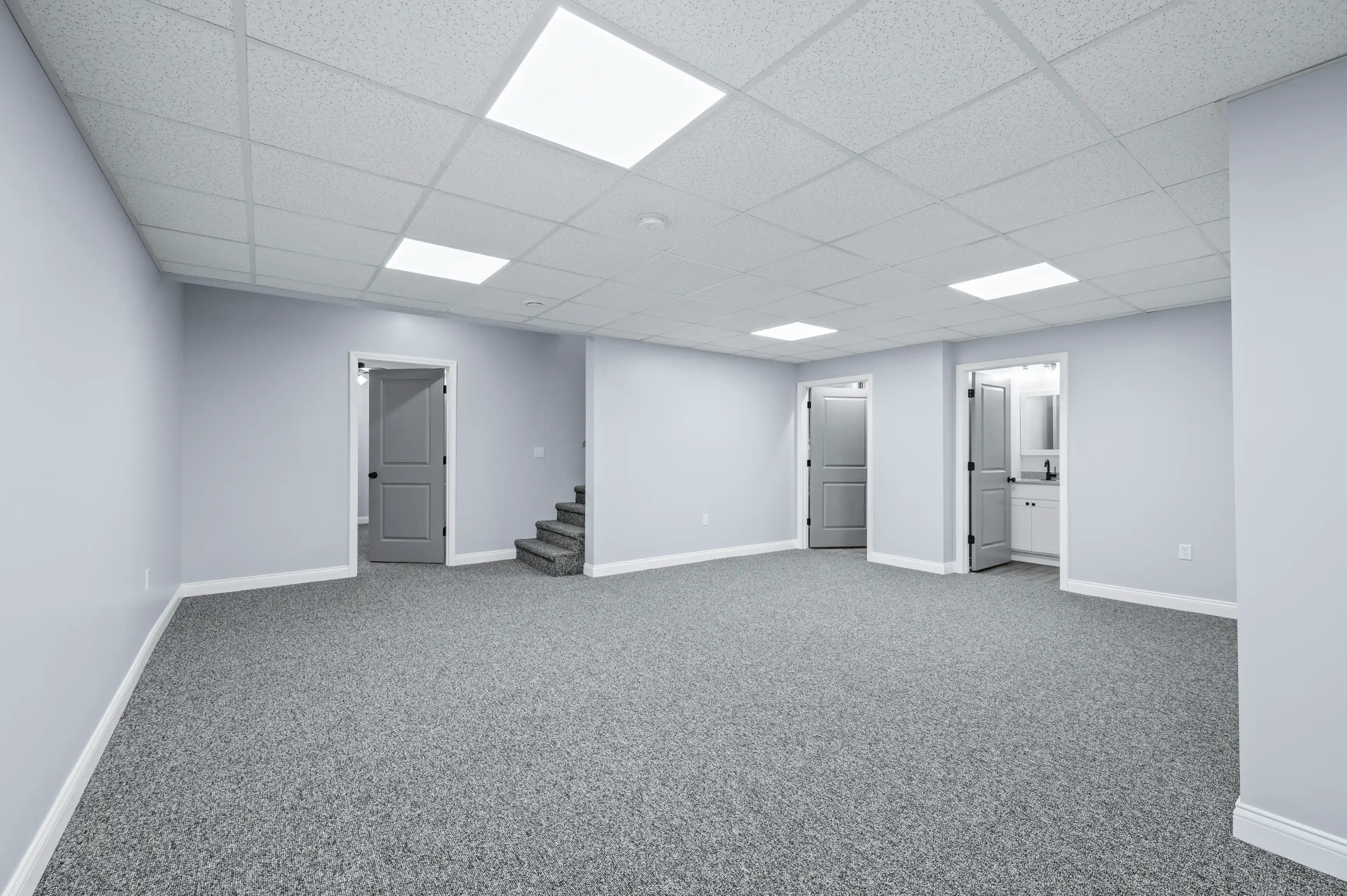 Spacious empty room with grey carpet, pale blue walls, recessed lighting, and multiple doors including one leading to a small kitchen area.