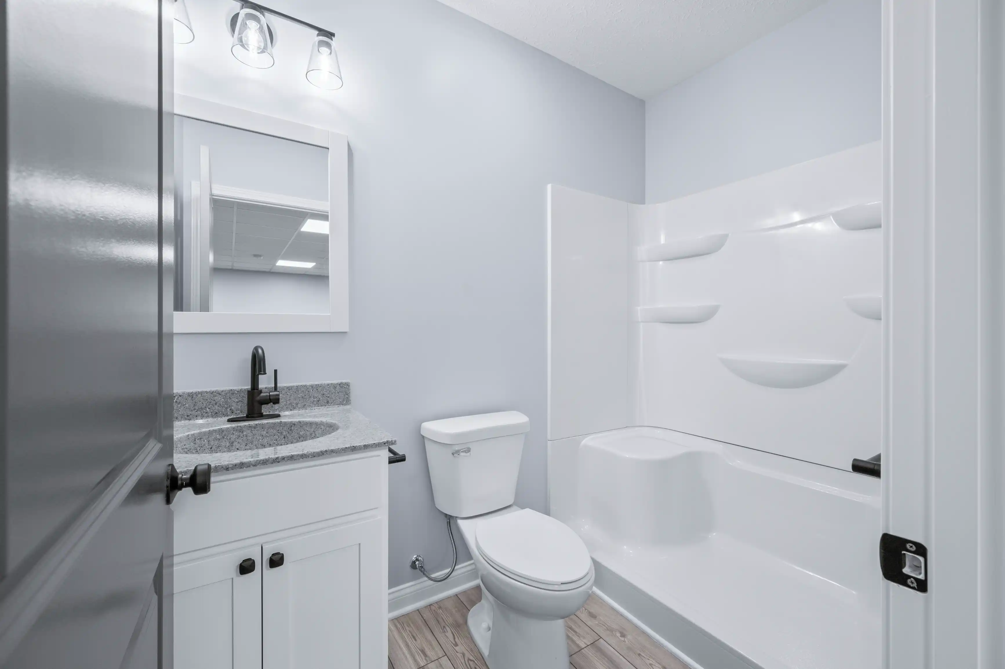 Modern bathroom interior with a white toilet, vanity cabinet with granite countertop and sink, a large mirror with overhead lights, and a built-in bathtub with shower area.