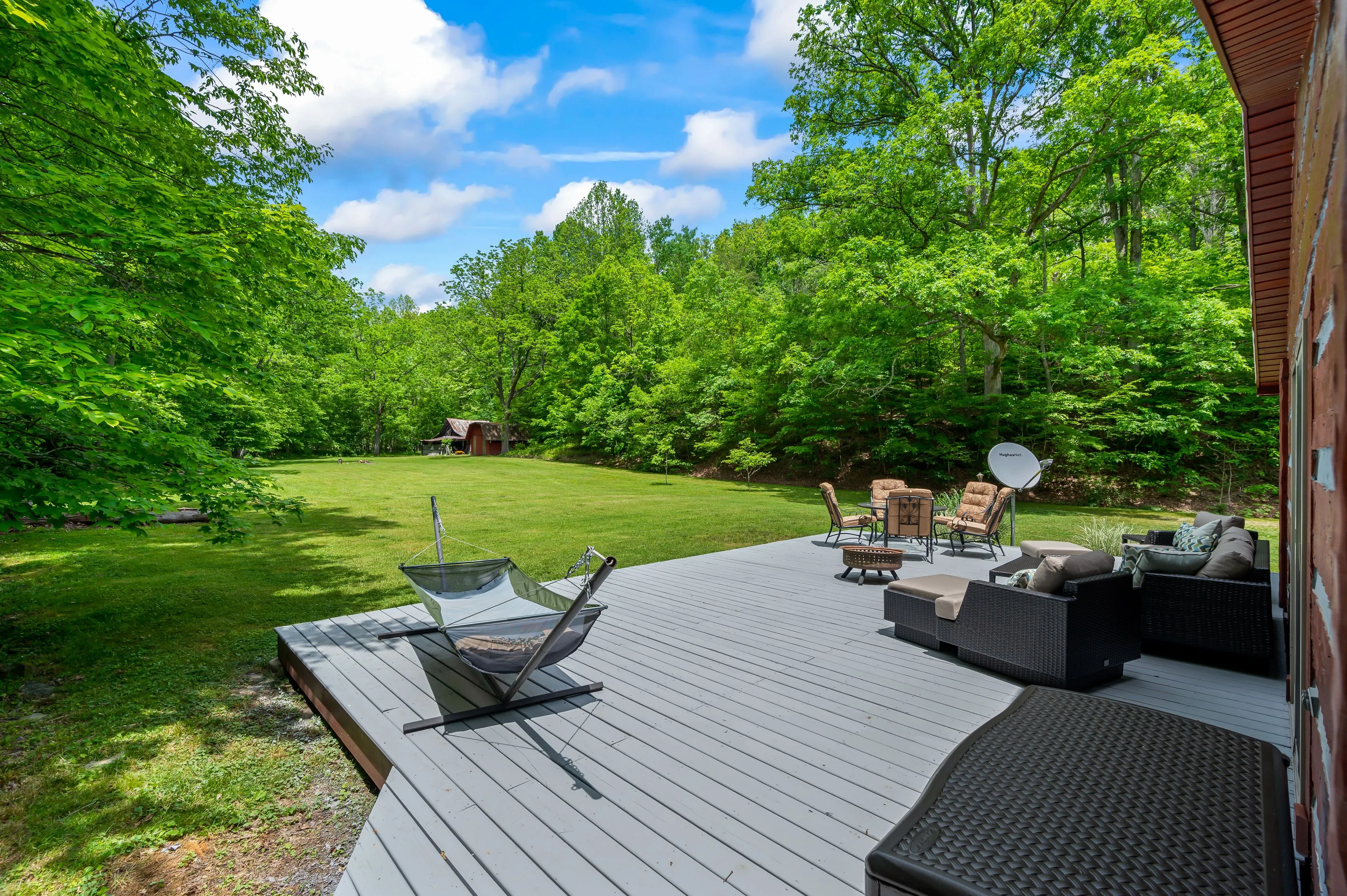 A spacious backyard with a large deck featuring outdoor furniture and a hammock, surrounded by lush green trees under a clear blue sky.