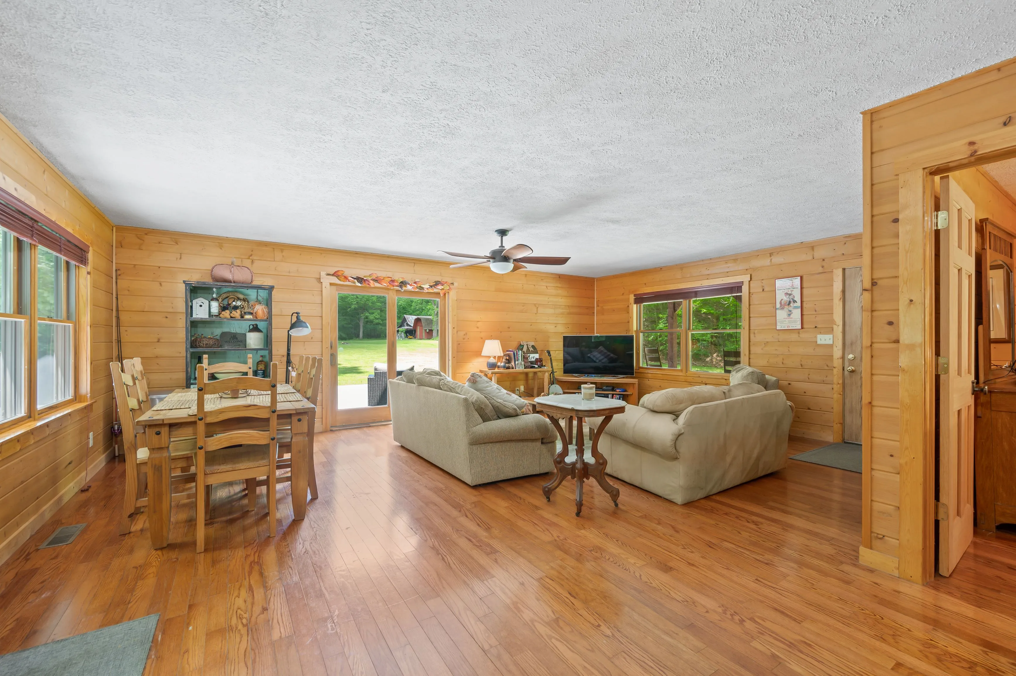 Wood-paneled living room with hardwood floors, comfortable seating, ceiling fan, and views of the yard through glass doors.