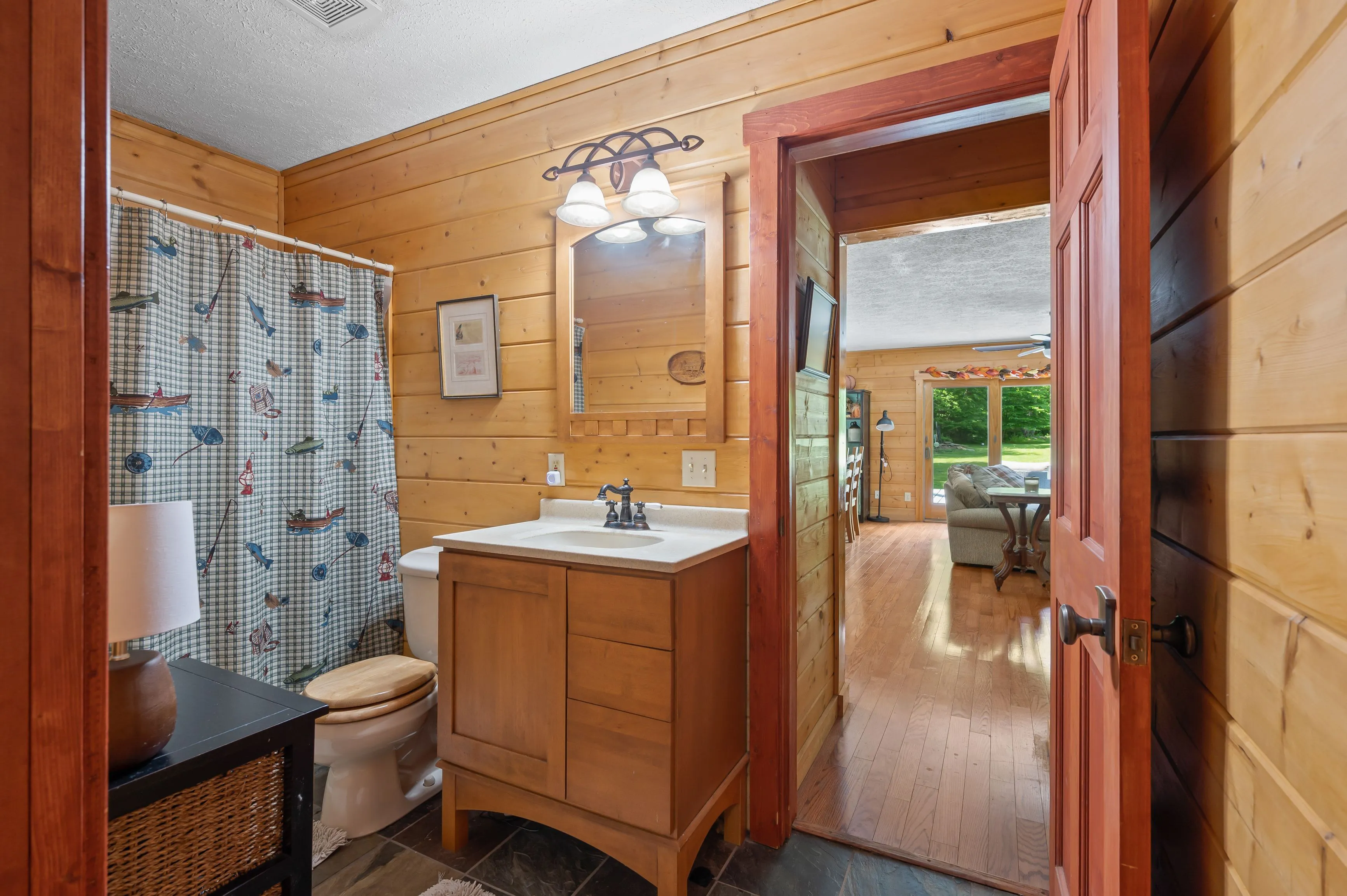 Interior of a rustic bathroom with wood paneling, showing a sink vanity, toilet, and shower curtain with a fishing motif, viewed through an open door leading to a living area.