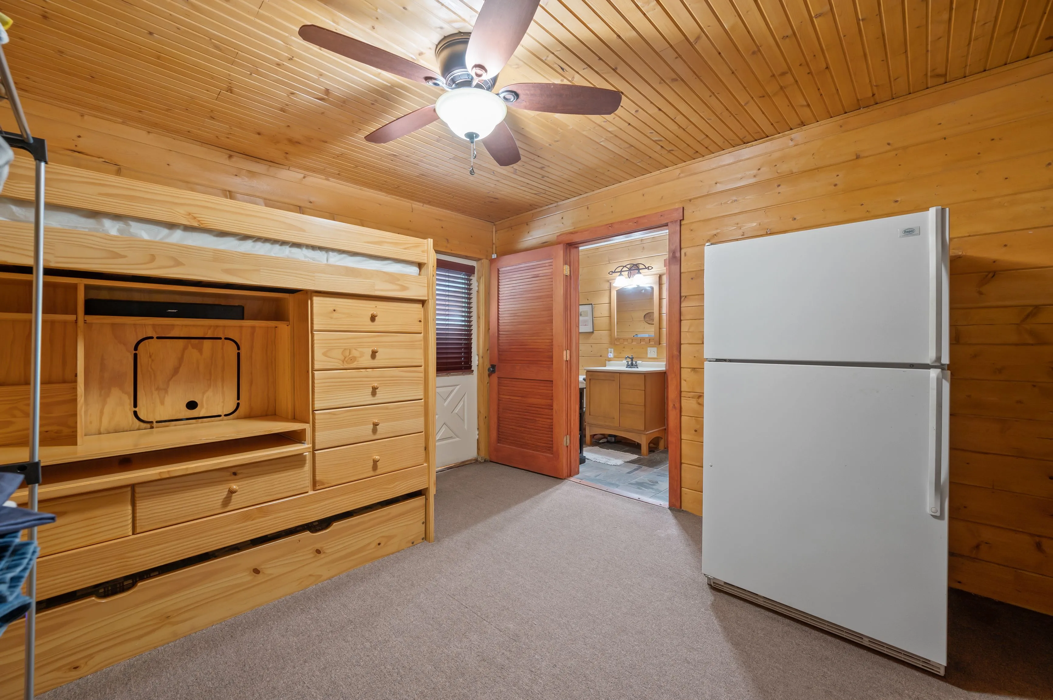 Interior of a wooden cabin room featuring a ceiling fan, built-in wooden cabinetry, a white refrigerator, and a door leading to a bathroom.