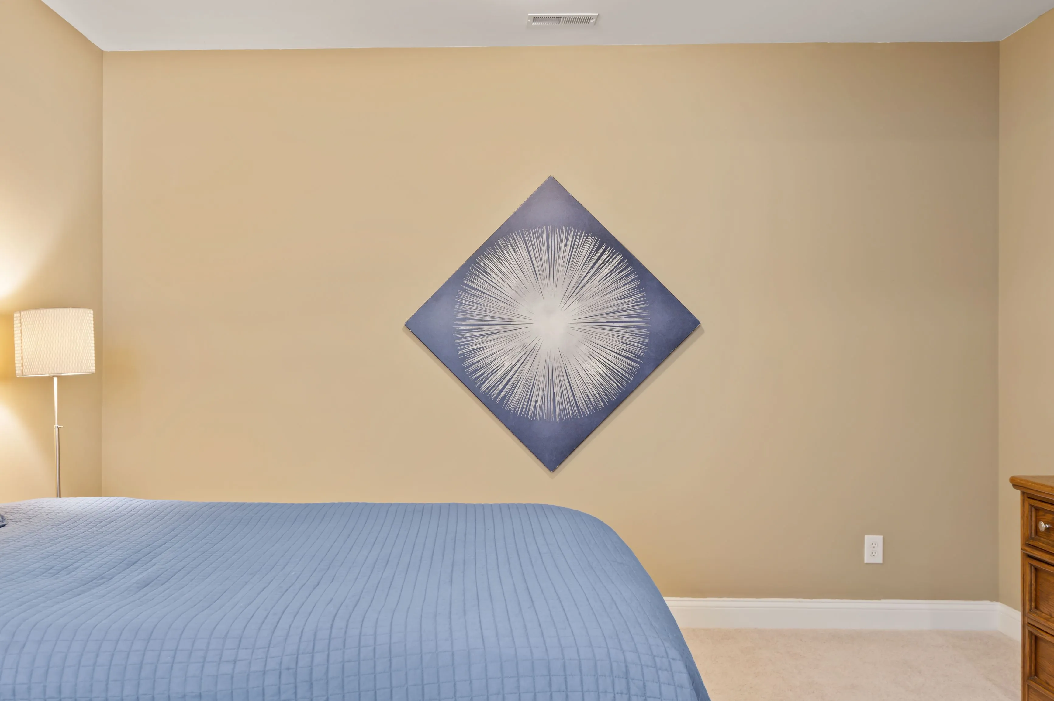 A tidy bedroom with a bed covered in a blue bedspread and an abstract piece of art hanging on the wall.