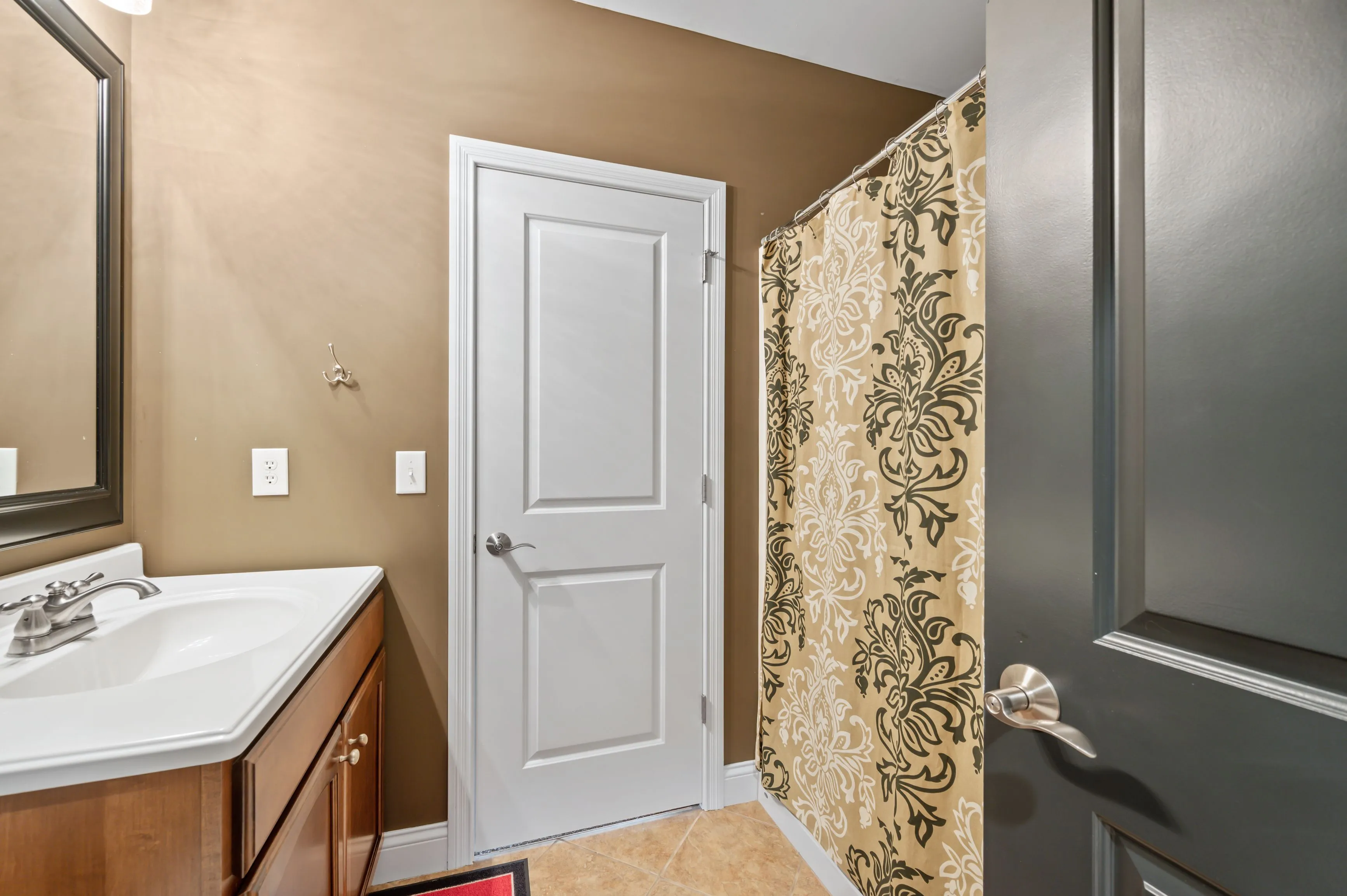 Interior of a modern bathroom featuring a white door, tan walls, a patterned shower curtain, a wooden vanity with a sink, and a gray towel hanging on the towel bar.