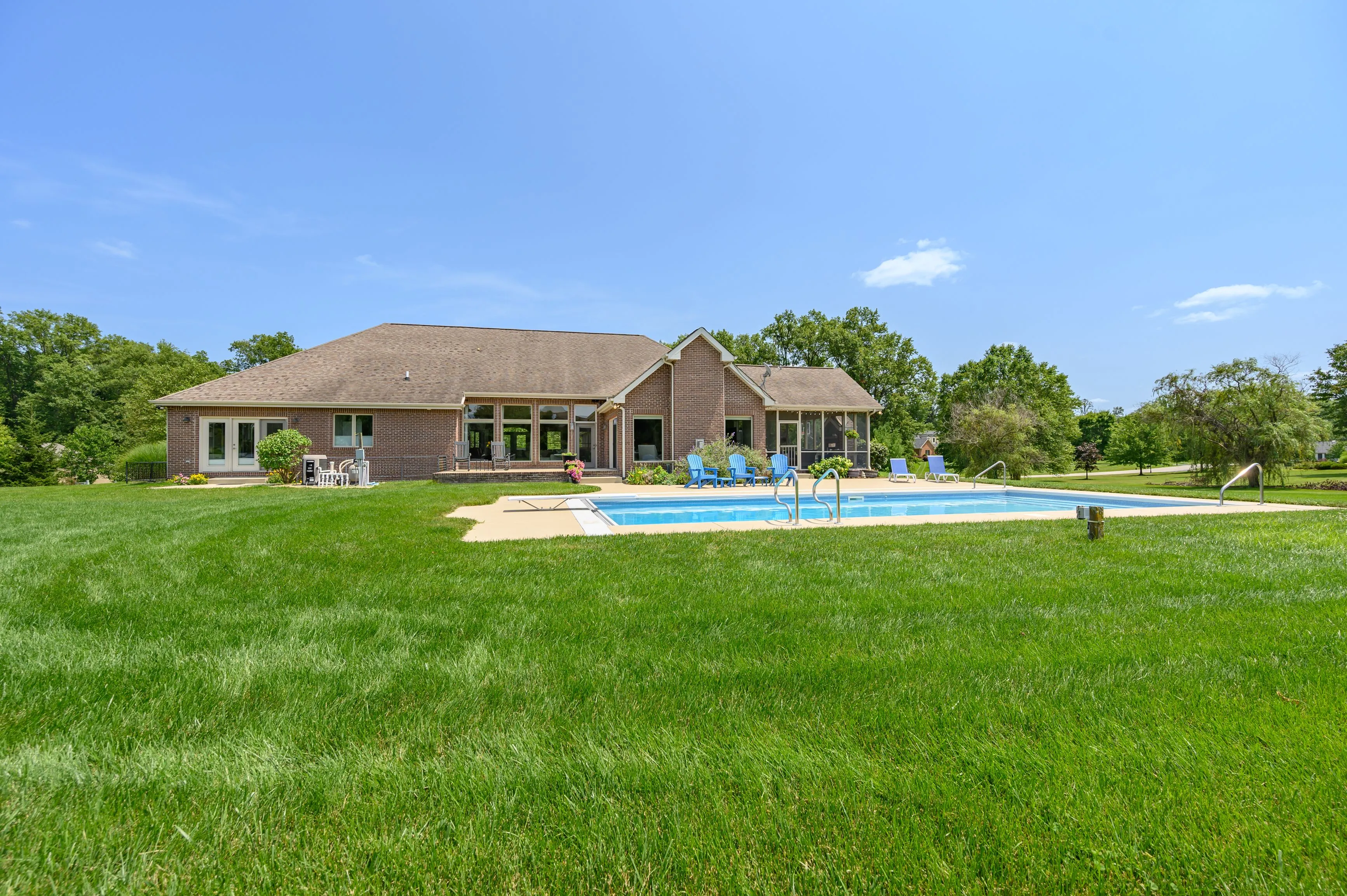 Single-story house with a shingled roof and an in-ground pool, surrounded by a large green lawn under a clear blue sky.