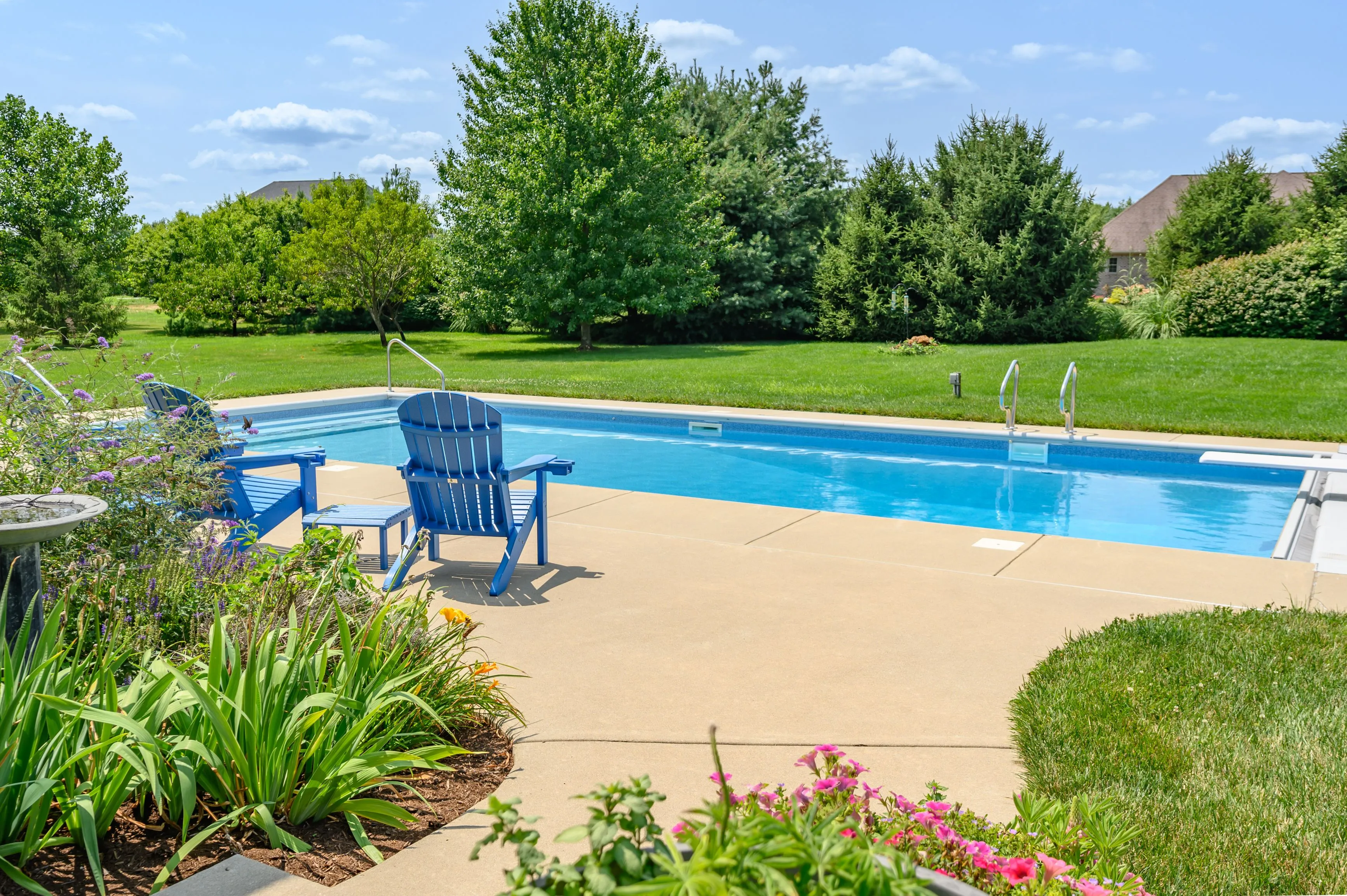 Backyard with an in-ground swimming pool, surrounded by a well-maintained lawn and garden, featuring a lone blue Adirondack chair on the poolside patio.