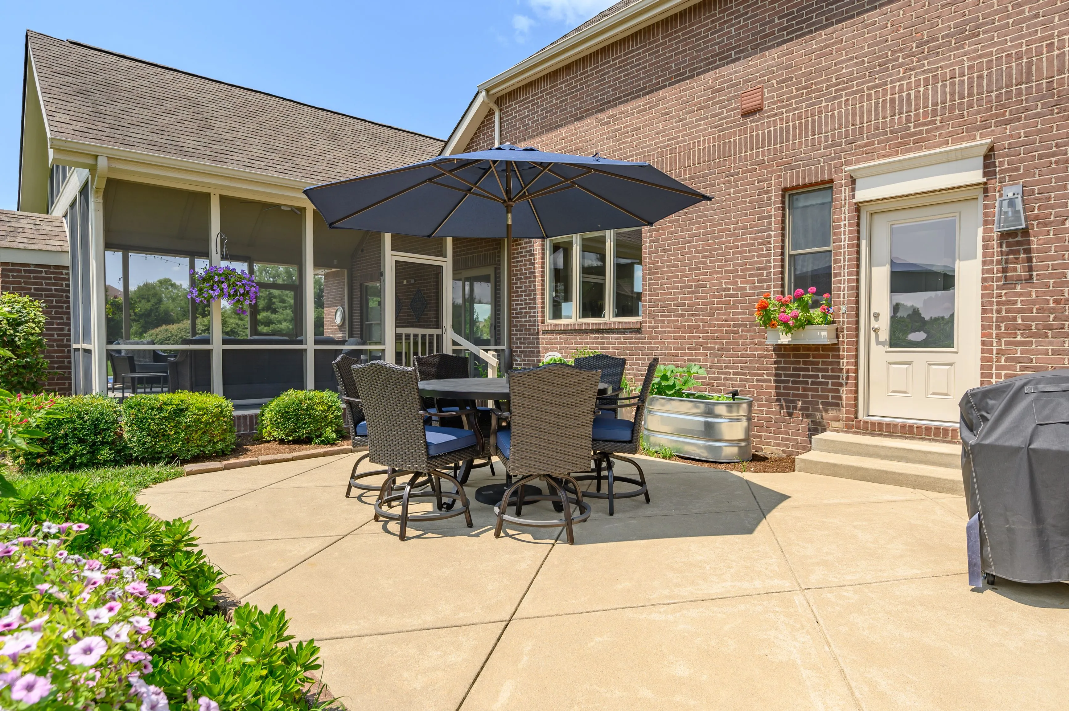Patio area with outdoor furniture including a table with chairs and an umbrella, adjacent to a brick house, with a clear sky above.
