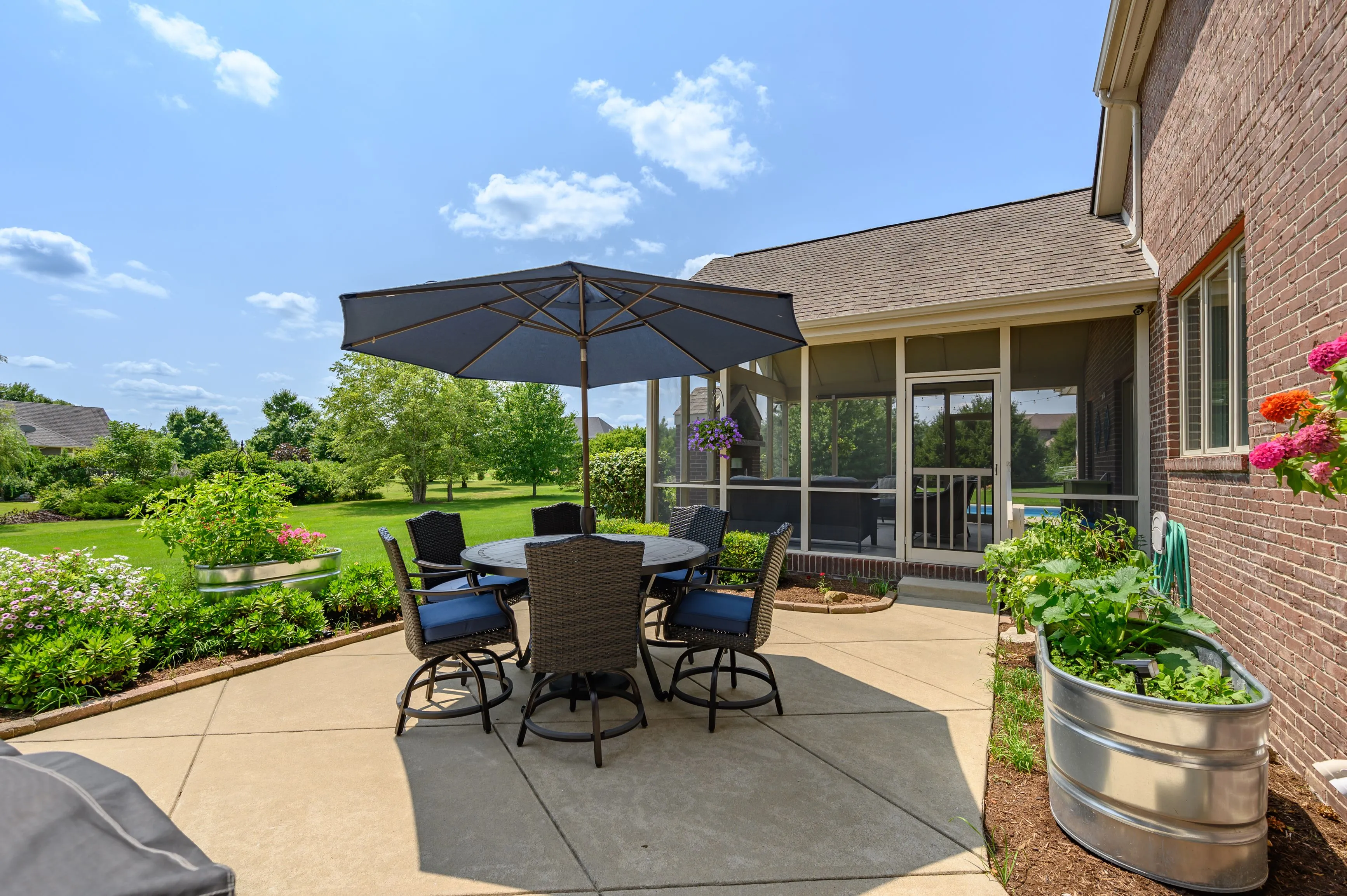 Outdoor patio area with dining table set under an umbrella, next to a brick house with a screened porch and a clear blue sky in the background.