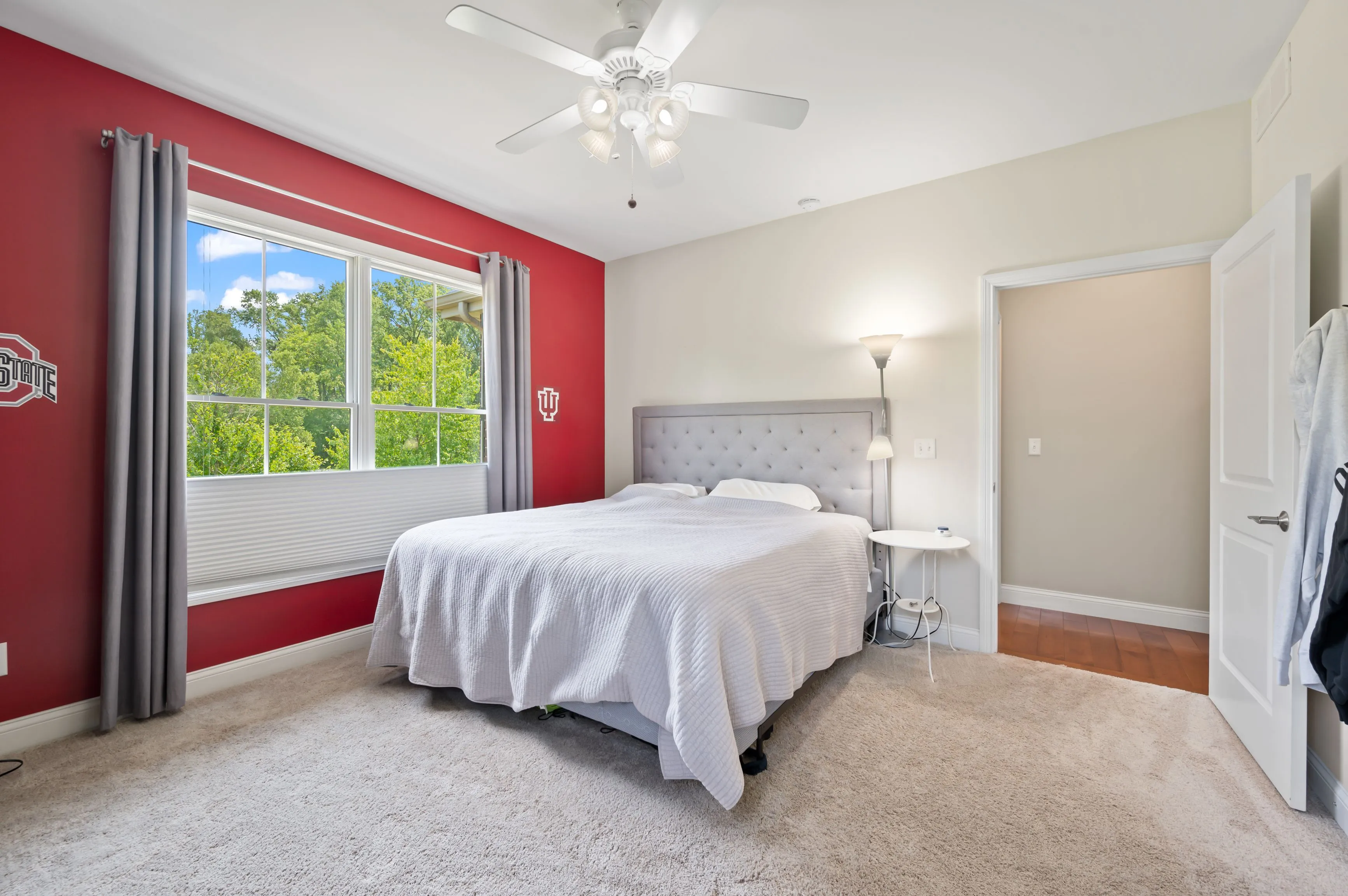 Bright bedroom with a large bed, red accent wall, window with a view of greenery, ceiling fan, and adjacent bathroom entrance.
