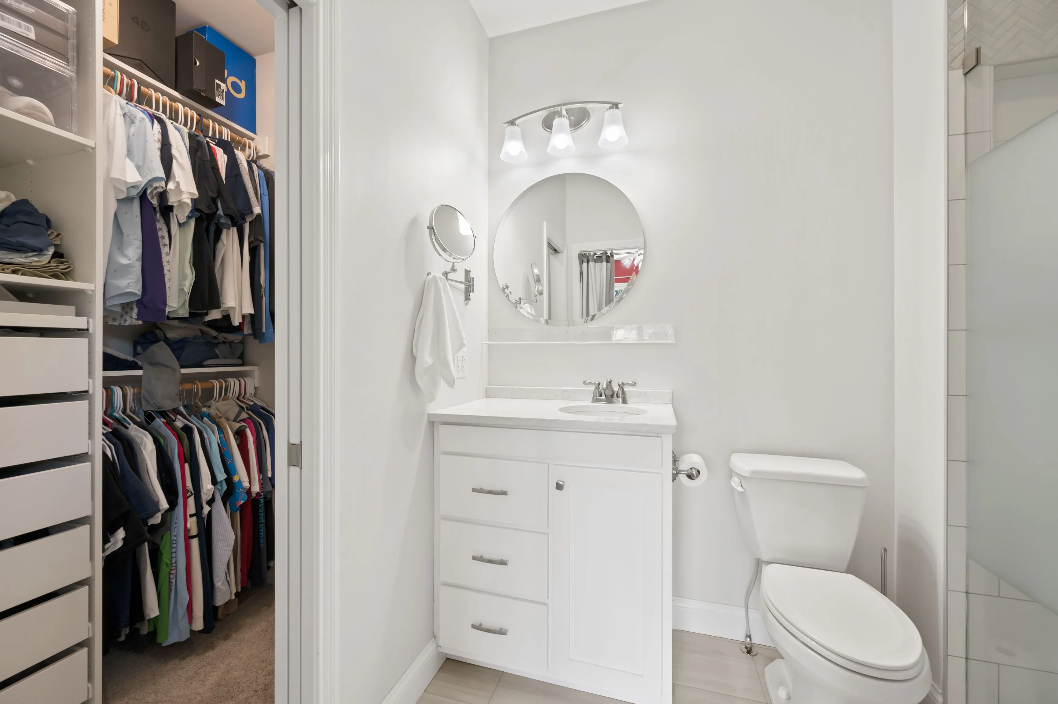 Interior view of a modern bathroom with a white vanity sink, mirror, and toilet, adjacent to a walking closet with shelves and clothes.